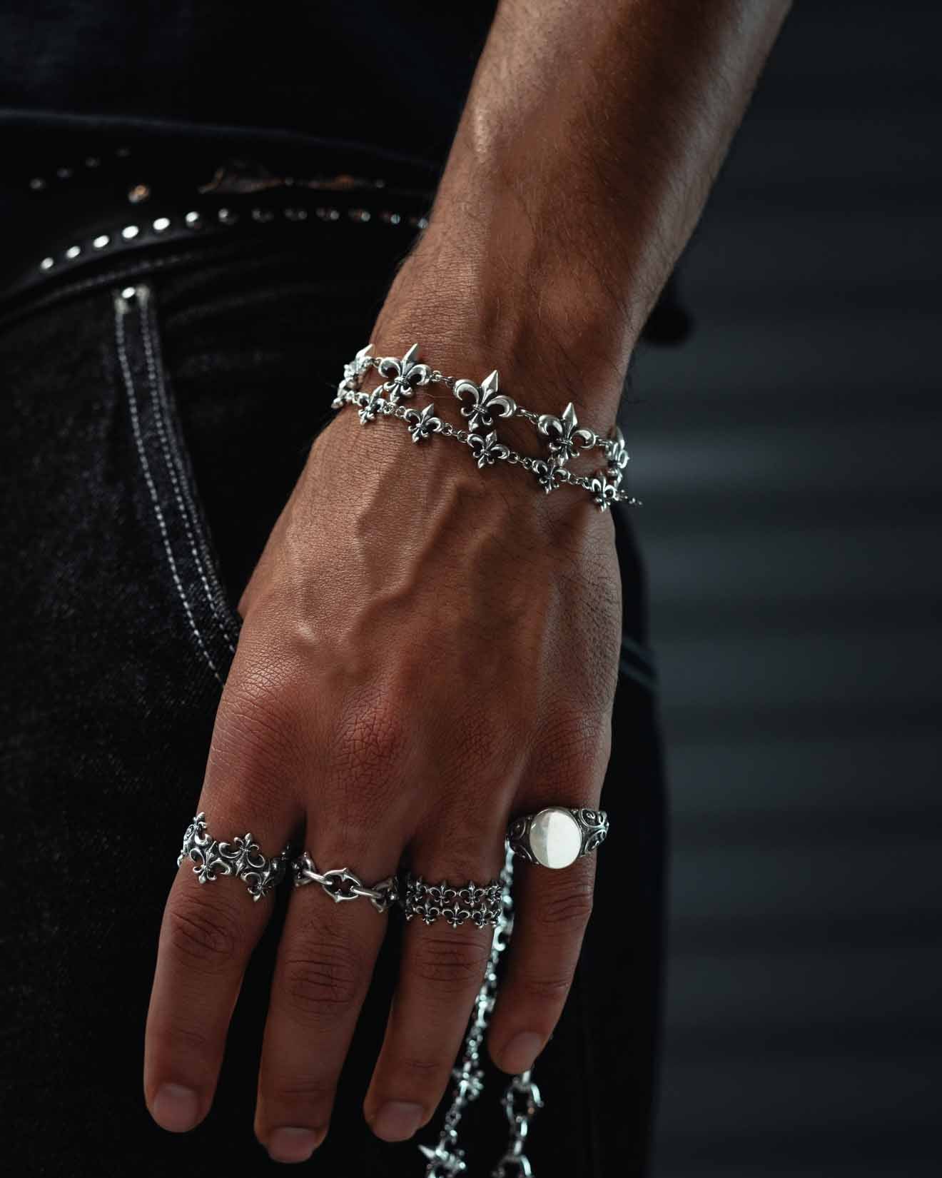 A close-up of a hand accessorized with multiple silver rings and the Statement Collective 925 Sterling Silver “Fleur De Lis” 15mm Bracelet, featuring ornate designs, resting against a black denim pocket with visible stitching.