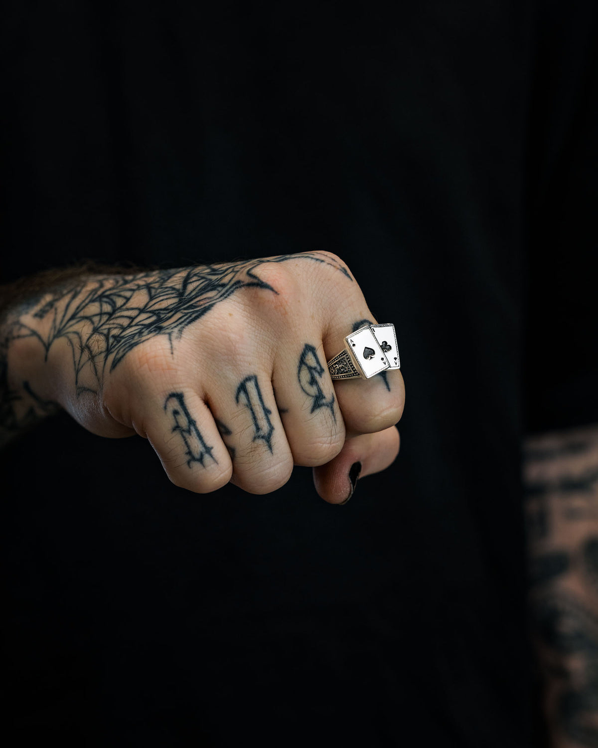 A tattooed hand with LIFE inked across the fingers displays the bold 925 Sterling Silver Pocket Aces Ring by Statement Collective, showcasing two playing cards—ace of hearts and ace of spades—in striking detail. A spiderweb tattoo adorns the wrist, while a dark background enhances the mysterious allure of this unique piece of macabre jewellery.