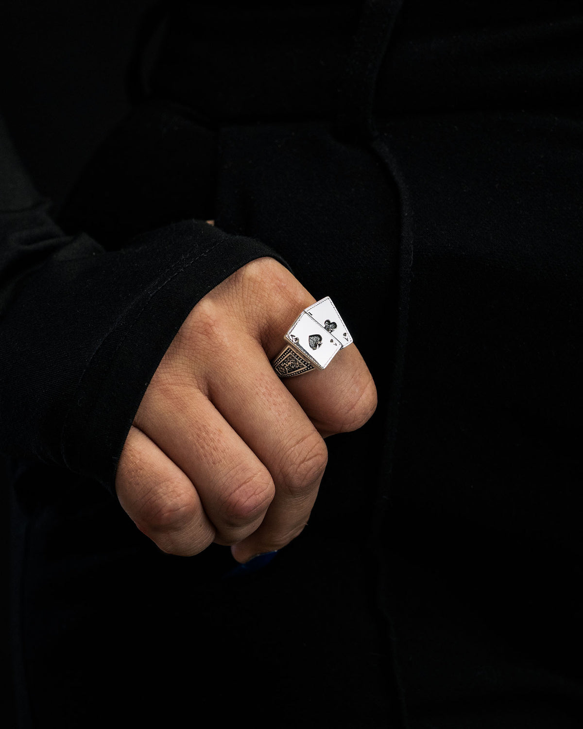 A hand adorned with the Statement Collective 925 Sterling Silver Pocket Aces Ring—crafted in the shape of a playing card ace—stands out against a black background and black clothing.