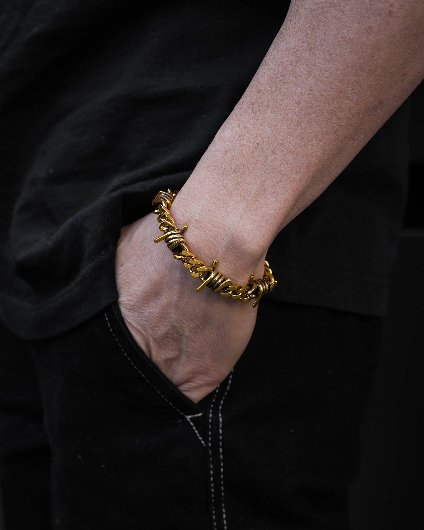 A person in a black shirt and pants has their hand in their pocket, showcasing the 9mm Barbed Wire Cuban Bracelet (Gold) on their wrist.