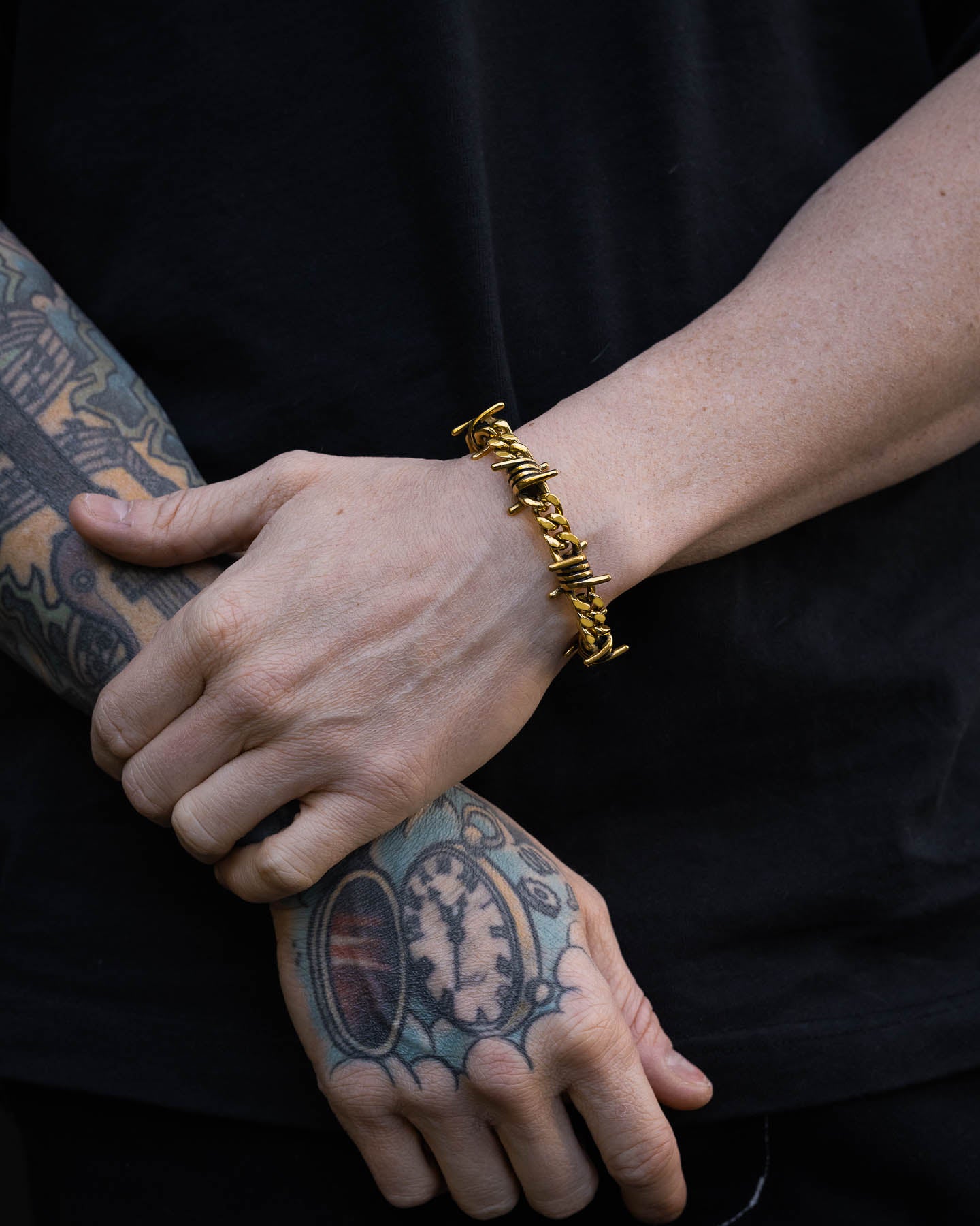 A person with tattooed hands wears the 9mm Barbed Wire Cuban Bracelet (Gold), standing against a black shirt background.