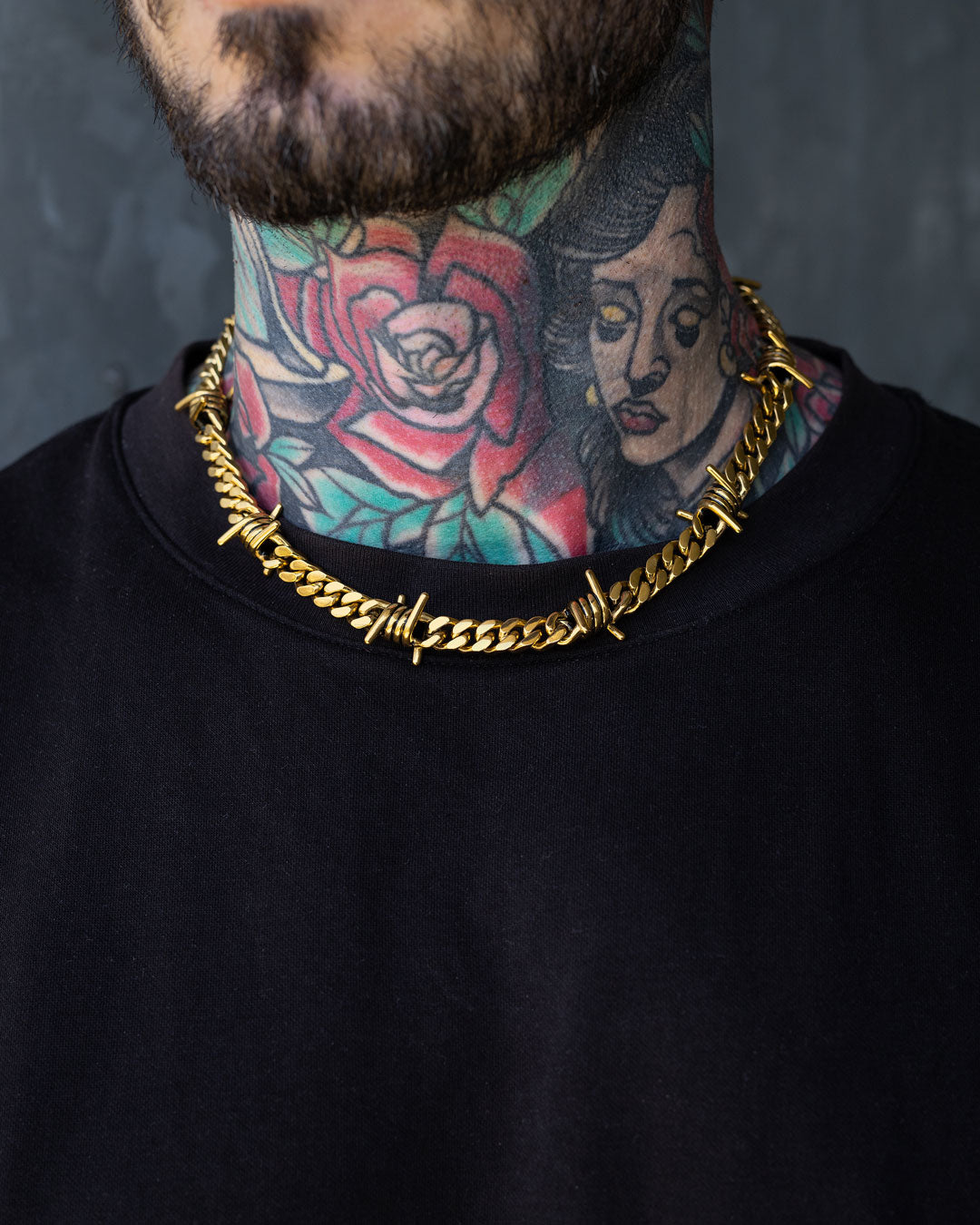 A person with colorful neck tattoos—a rose and a woman's face—wears the 9mm Barbed Wire Cuban Chain (Gold) over a black shirt.