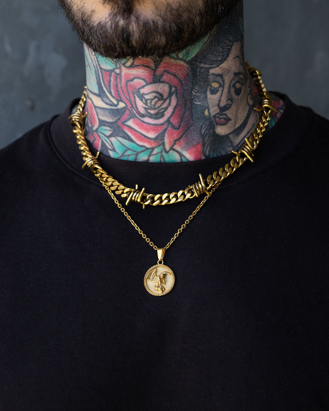 A close-up of a person’s neck wearing two gold necklaces, including the 9mm Barbed Wire Cuban Chain (Gold) and a pendant, with colorful rose and portrait tattoos visible on their neck.