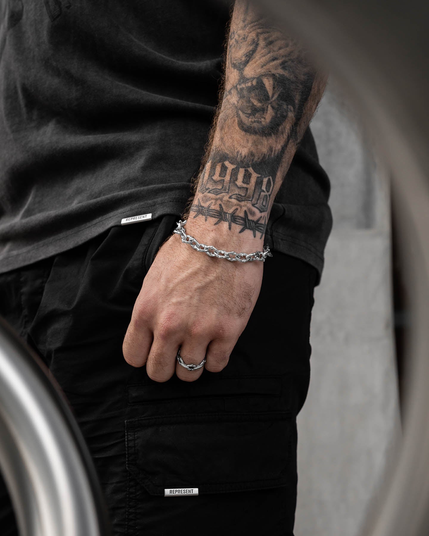 A close-up of a tattooed arm and hand wearing "The Cathedral" 9mm Spiked Bracelet, styled with tiger, 1998, and barbed wire tattoos. They wear a black shirt and cargo shorts.