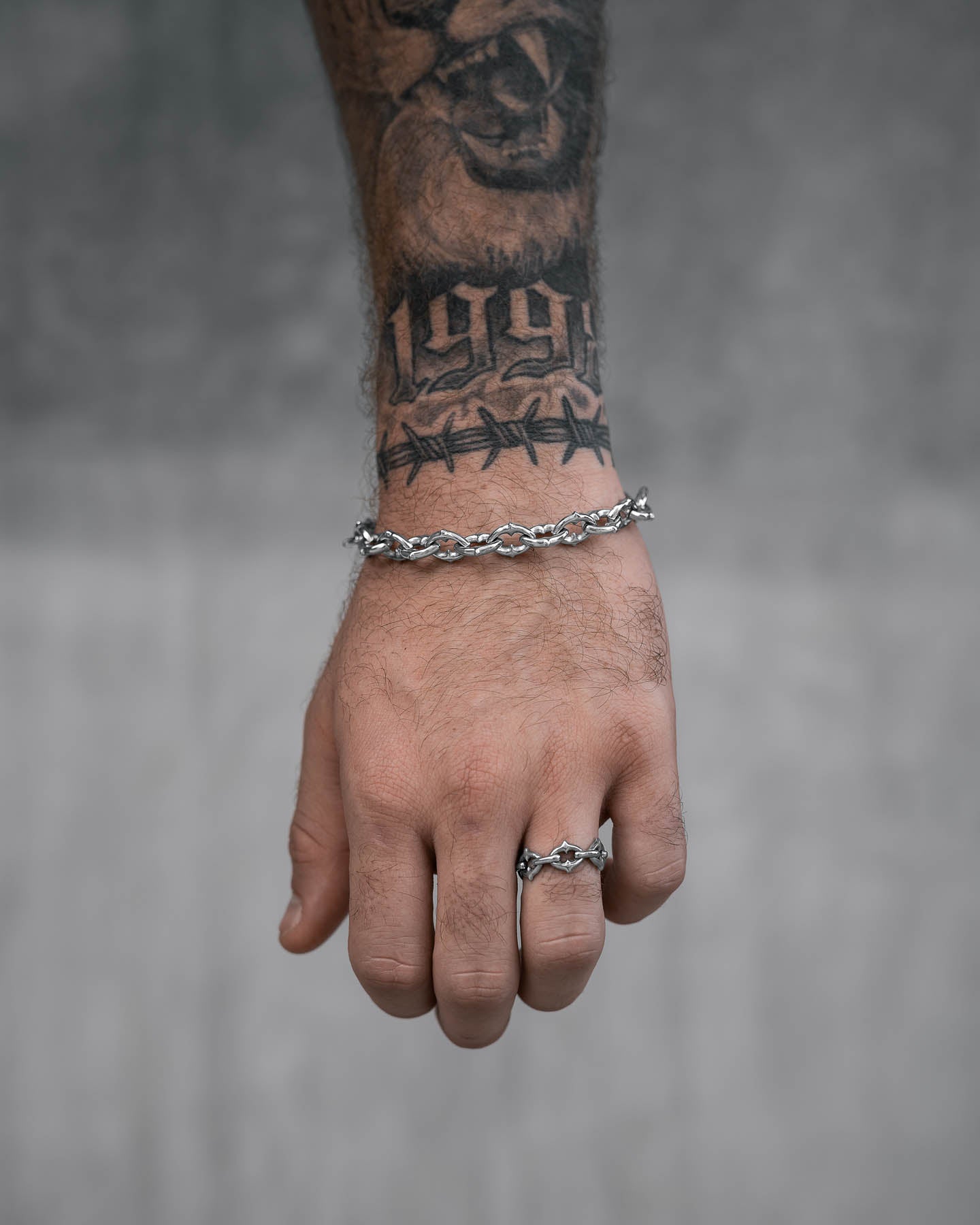 A tattooed hand with a “1994” barbed wire tattoo on the wrist wears "The Cathedral" 9mm Spiked Bracelet, non-tarnish and waterproof, against a blurred gray background.