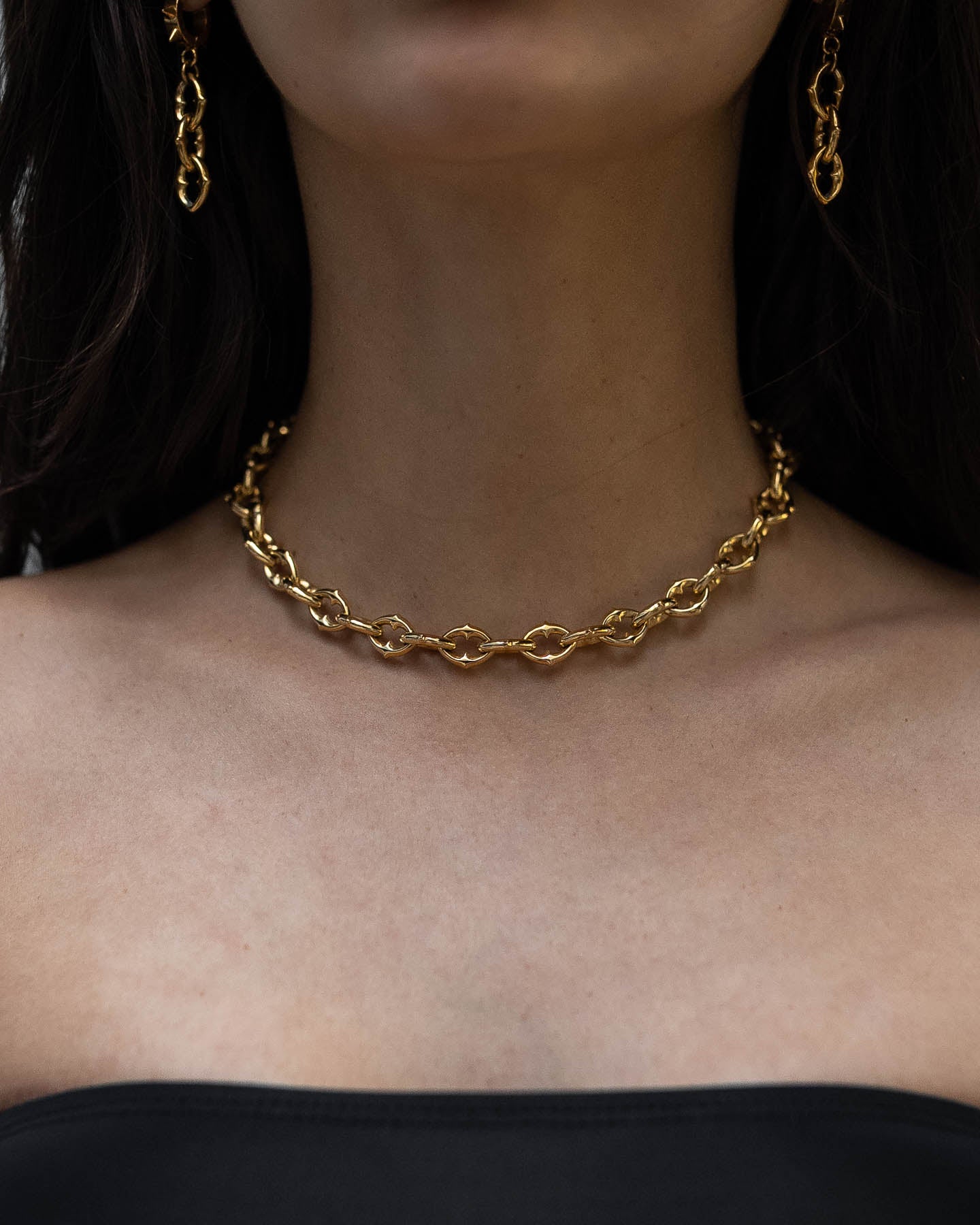 A woman wears a strapless black top and "The Cathedral" 9mm Spiked Chain (Gold) necklace, with matching gold chain earrings. The image highlights her neck and shoulders.