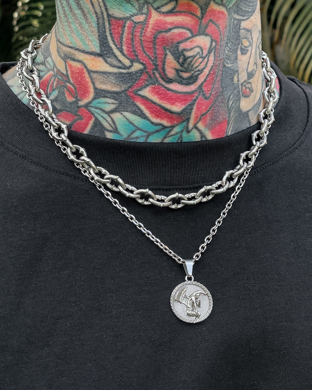 A person with colorful neck tattoos, including a red rose and a woman's face, wears "The Cathedral" 9mm Spiked Chain and a thin silver necklace with a round pendant over a black shirt.