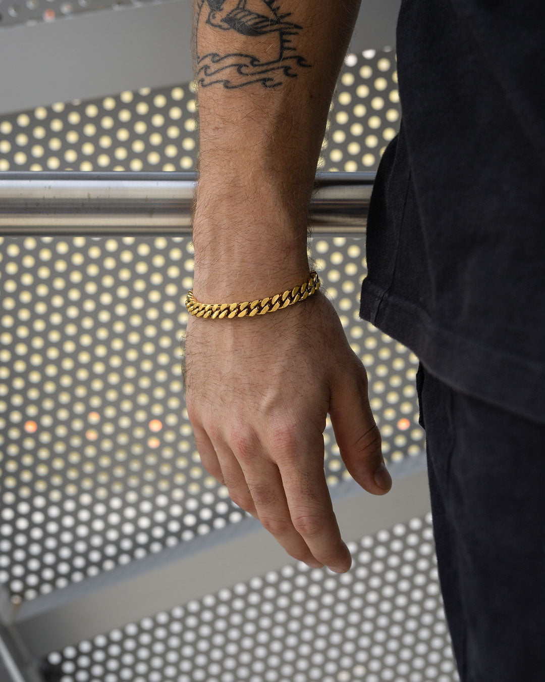 A person with a bird tattoo on their arm and hand wears the striking 9mm Cuban Bracelet (Gold), standing by a metal railing against a perforated metal backdrop, dressed in black.
