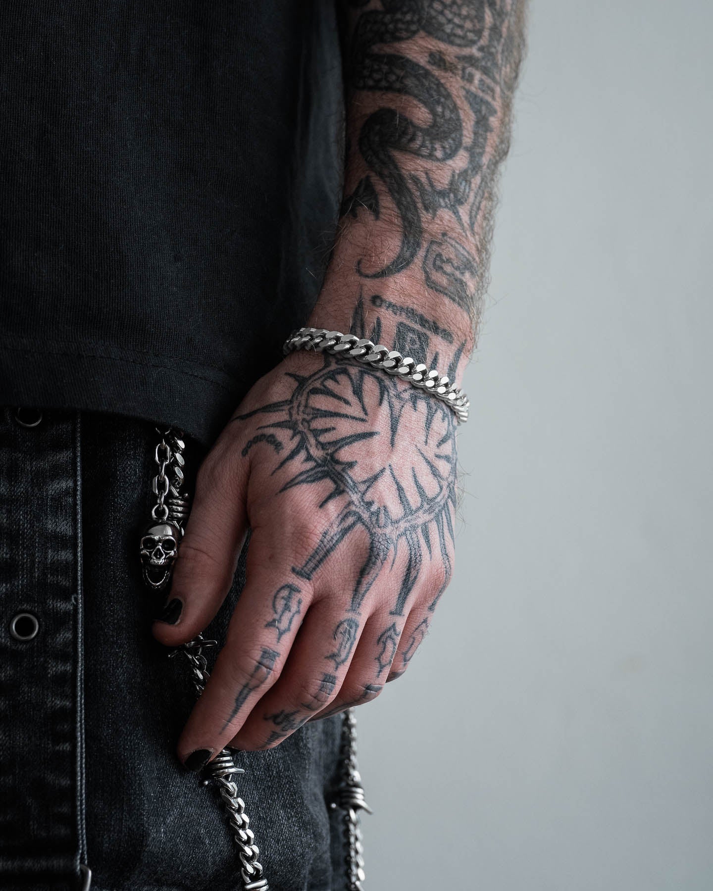 A close-up of a tattooed hand with black nail polish wearing the 9mm Cuban Bracelet. The person is dressed in dark clothing, and a skull chain accessory is attached to their pants.
