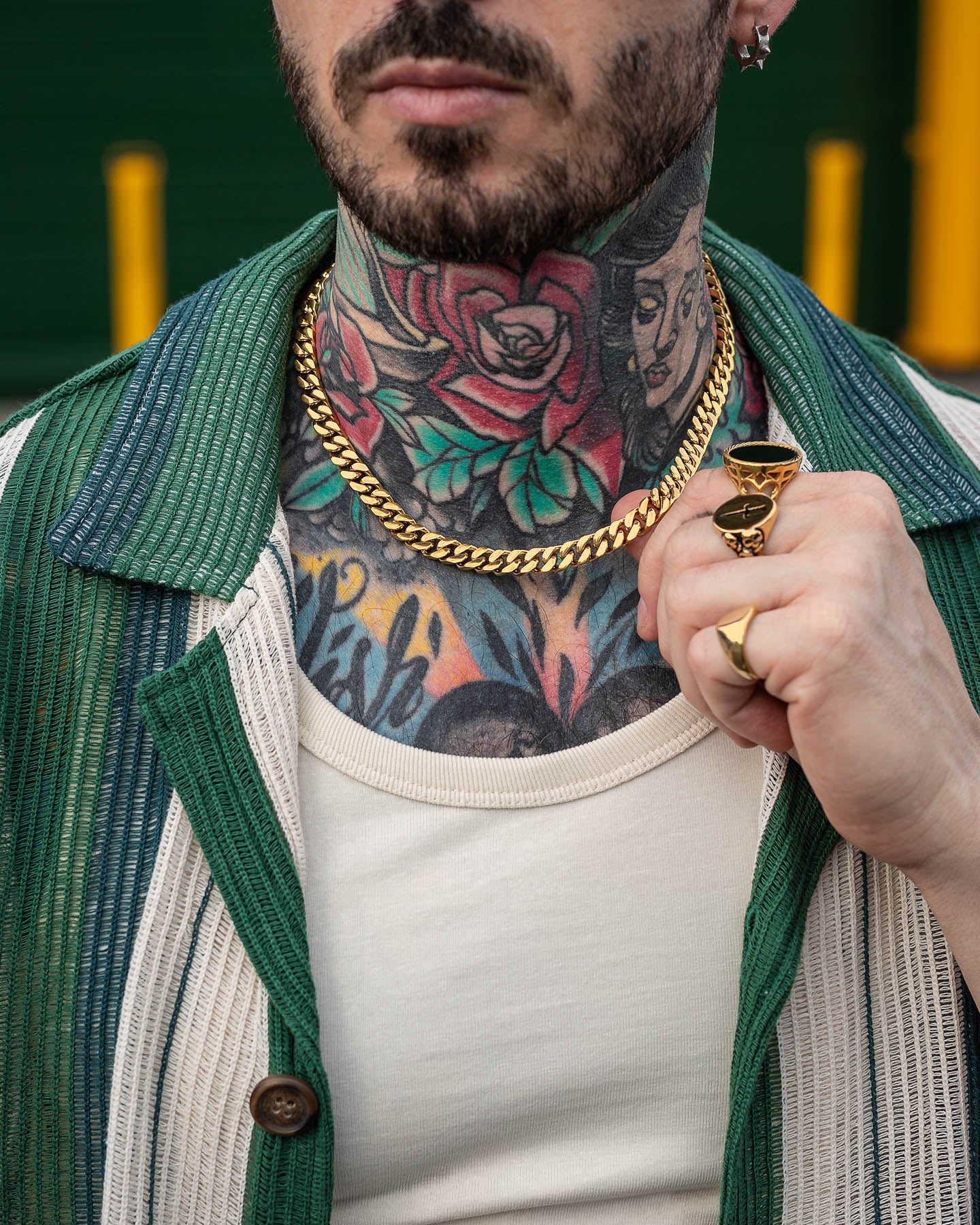 A person with colorful neck tattoos wears the 9mm Cuban Chain (Gold), layered rings, a white shirt, and a green-striped jacket, holding their collar with one hand.