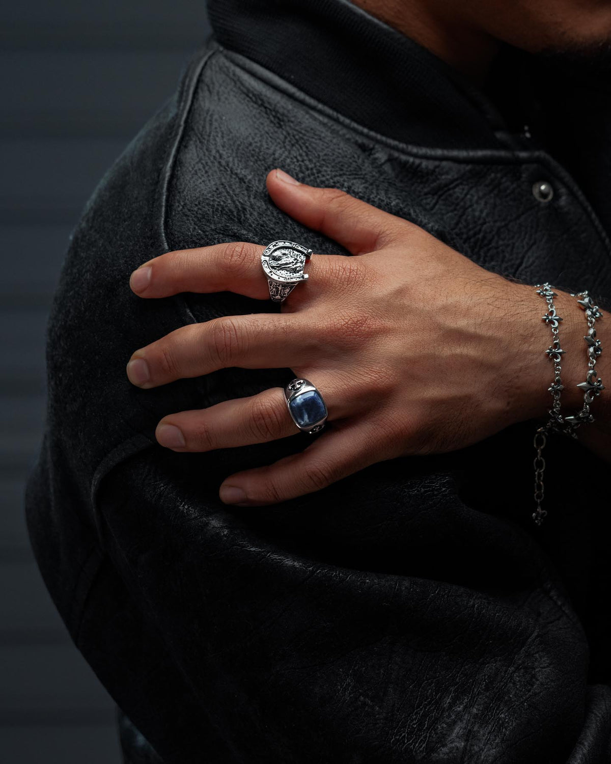 A person in a black leather jacket rests one hand on their shoulder, showcasing two rings—including the Statement Collective Sodalite Stone Ring with its striking blue stone—and a silver beaded bracelet.