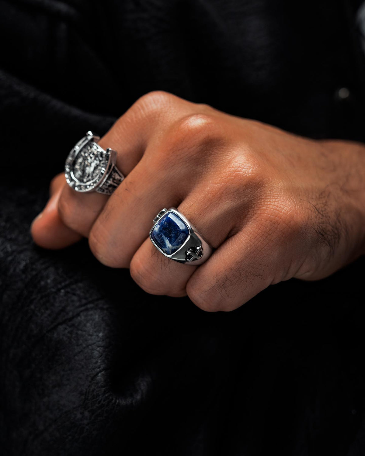 A hand adorned with two silver rings: one is the Statement Collective Sodalite Stone Ring, crafted from stainless steel and featuring a striking blue rectangular stone with a star detail, while the other showcases an ornate, circular design inspired by graveyard jewelry. The background is dark and textured.