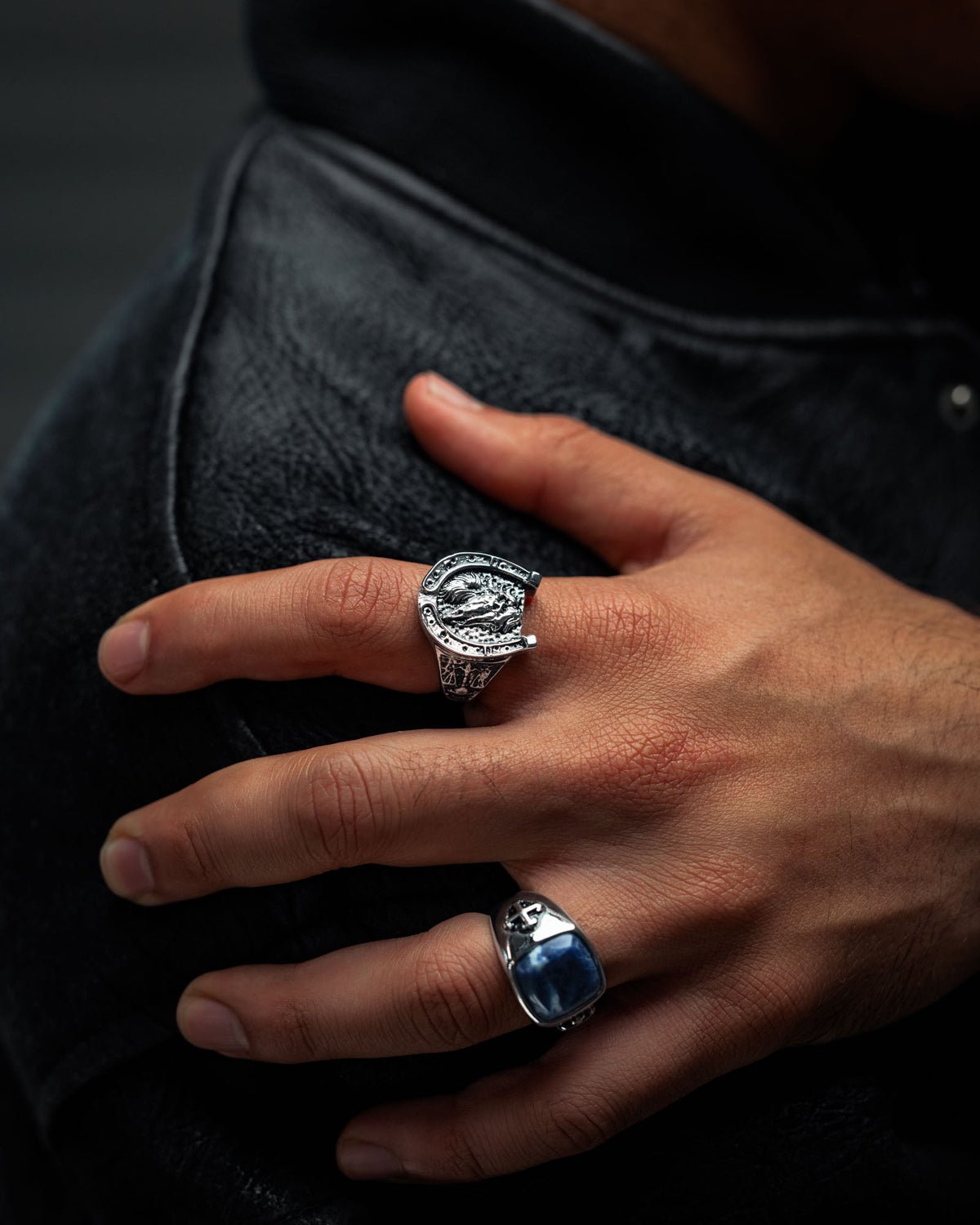 A hand adorned with two silver rings—one being Statement Collective’s Bronco Ring, notable for its intricate detailing, and another featuring a striking blue stone in a graveyard motif—rests gently on a black leather jacket, offering a refined nod to macabre jewellery.