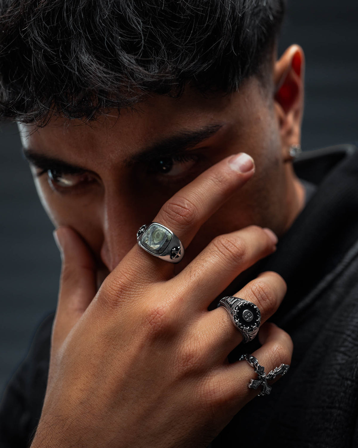 A person with short dark hair gazes intensely, partially covering their face with their hand. Their fingers are adorned with several bold silver rings, including the striking Kambaba Jasper Stone Ring from Statement Collective, which adds a touch of mysterious elegance to their jewellery ensemble. An earring complements the look, while a dark textured jacket and shadowy background further highlight their enigmatic style.