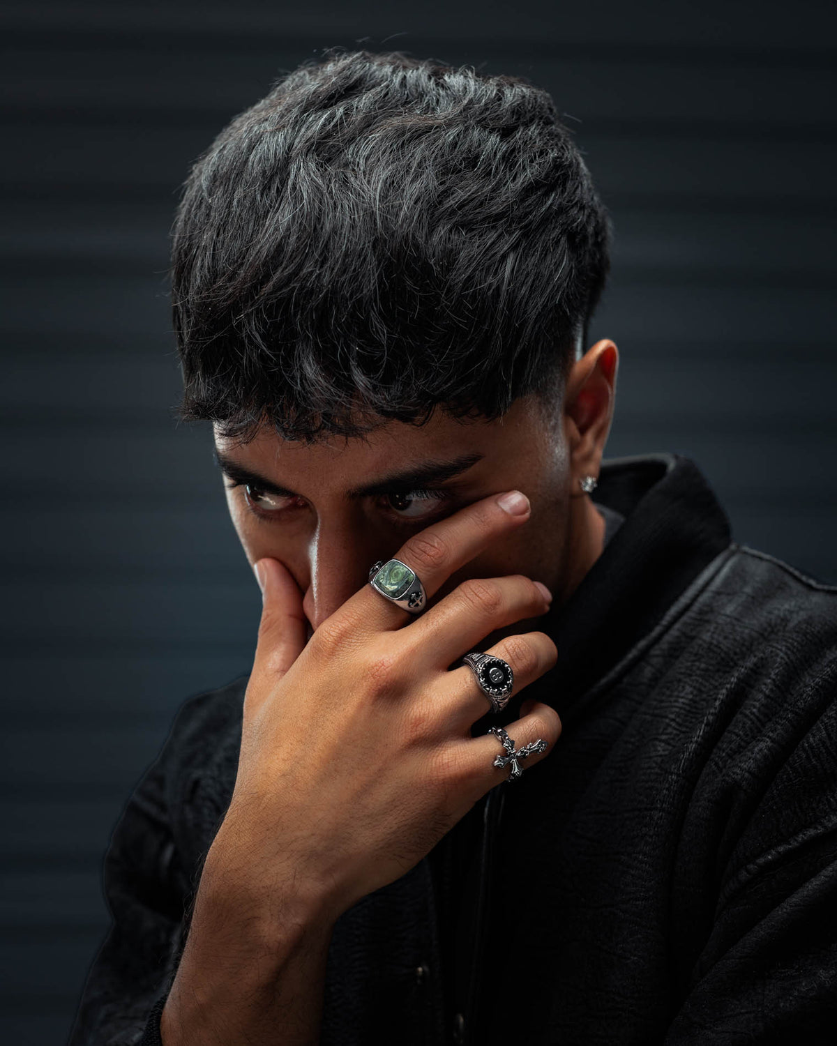 A person with short dark hair partially covers their face with their hand, showcasing several rings—the standout being the Statement Collective Kambaba Jasper Stone Ring. They are dressed in a black textured jacket and glance to the side against a softly blurred dark background.