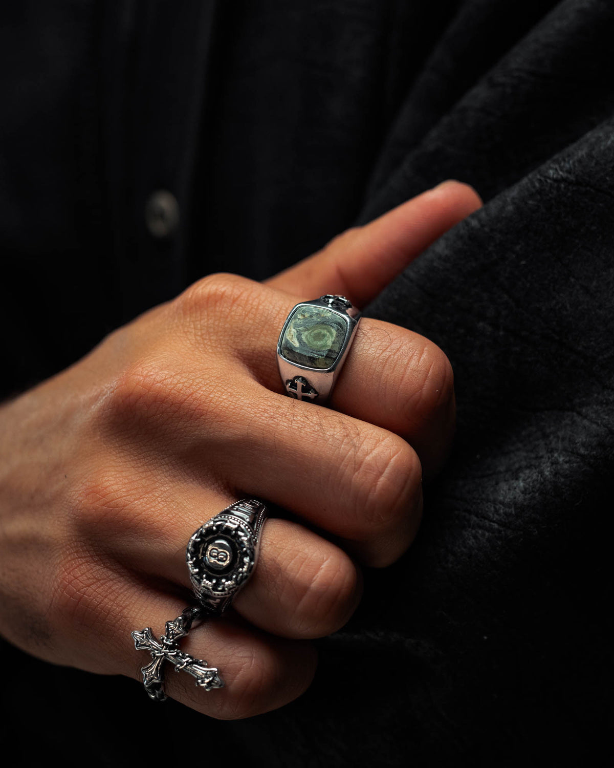 A hand wearing two silver rings from Statement Collective, one being the Kambaba Jasper Stone Ring set with a striking green gemstone and the other showcasing a cross design inspired by macabre jewellery, displayed against a dark textured background.