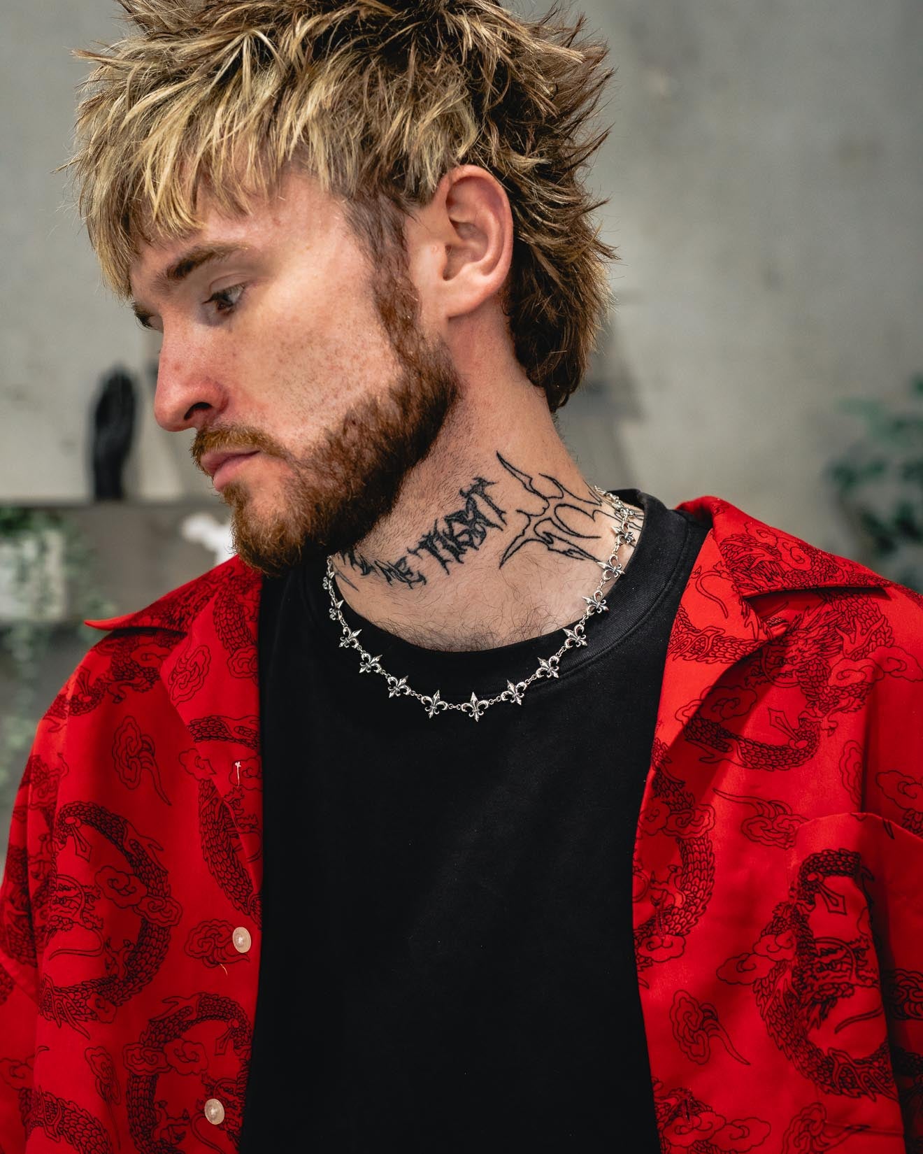 A person with blonde, messy hair and a beard wears a red patterned shirt layered over a black t-shirt. Around their neck is the Statement Collective 925 Sterling Silver “Fleur De Lis” 15mm Chain. A large tattoo covers their neck, and they look down with a neutral expression.