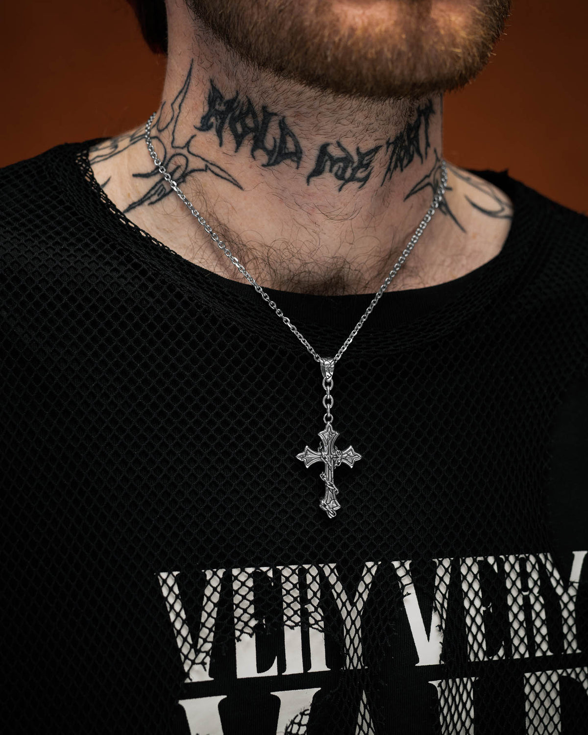 A person with a reddish-brown beard wears Statement Collective’s Mini “The Ruins” Cross Pendant necklace, a black mesh shirt, and has black script tattoos on their neck.