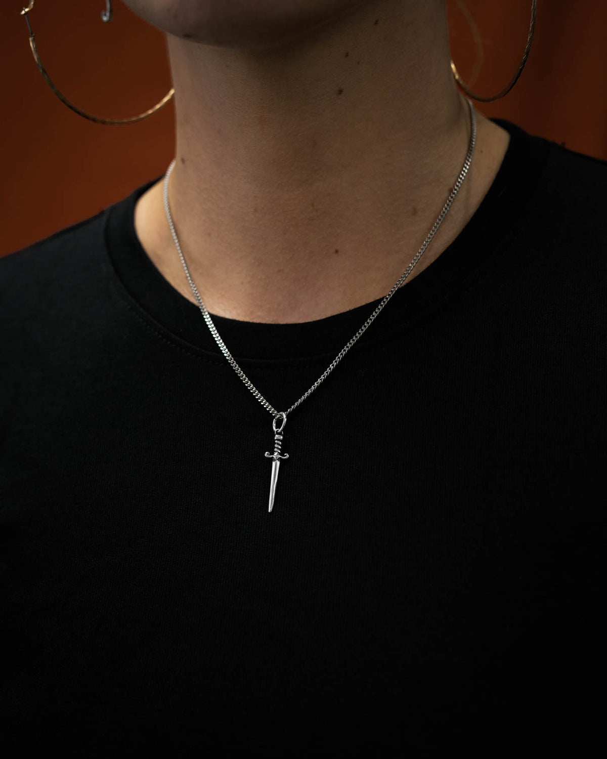 Person wearing a silver necklace with a mini dagger pendant on a dark orange background