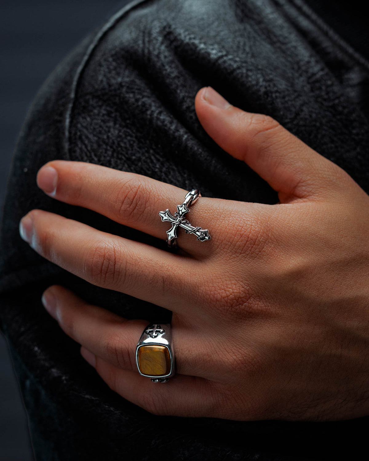 A hand with two rings—one, Statement Collective’s “The Ruins” Chained Cross Ring in silver, the other a macabre piece set with a square yellow stone—rests on a black leather surface.