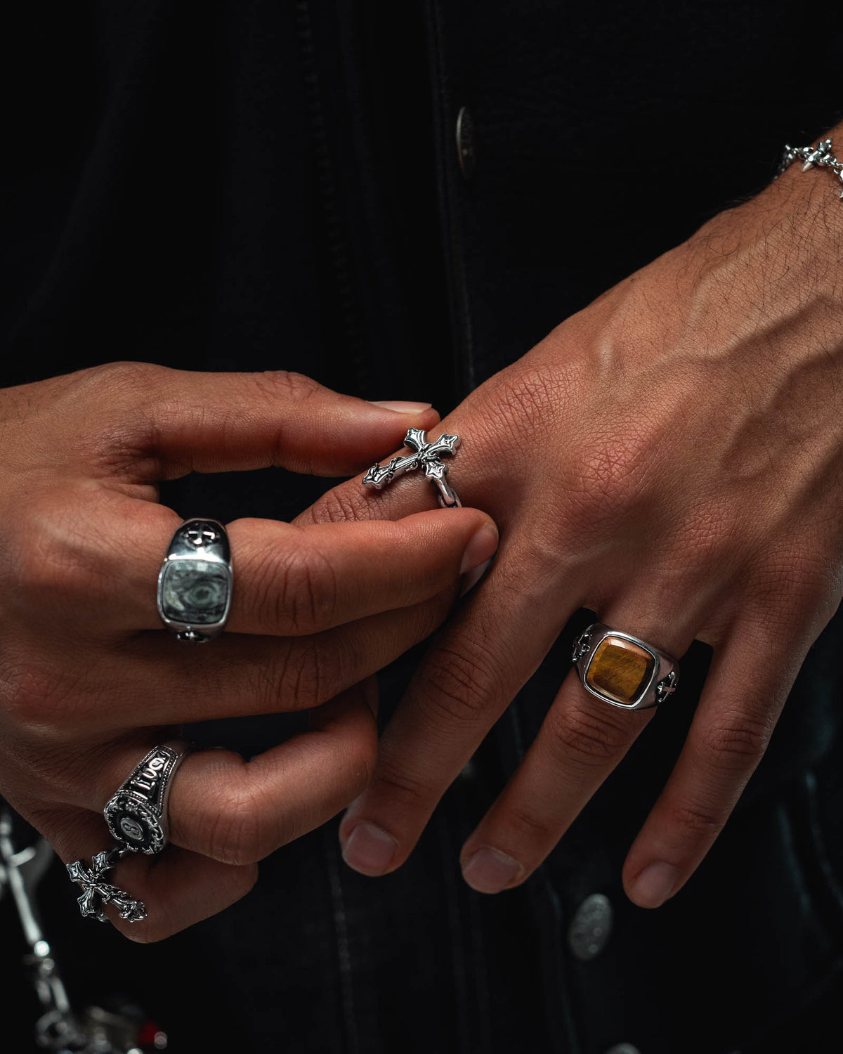 Close-up of two hands adorned with several silver rings from Statement Collective, including “The Ruins” Chained Cross Ring—a design inspired by macabre jewelry. The dark background accentuates the intricate details of the rings and the rich texture of the skin.