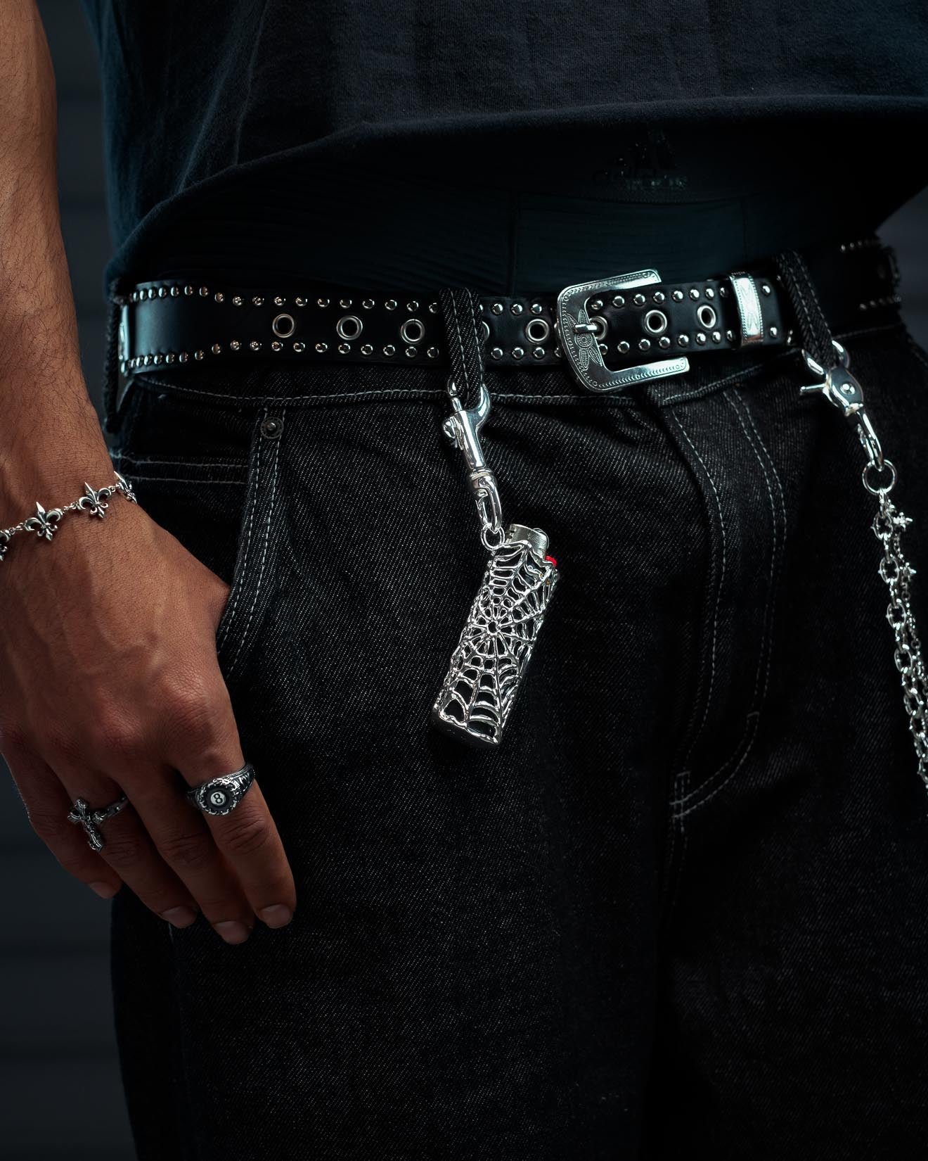 A person wearing black jeans, a studded belt, and silver accessories—including rings, a chain bracelet, and the Statement Collective Spiderweb Lighter Sleeve for their Bic lighter—has it hanging alongside a spiderweb keychain from their belt.