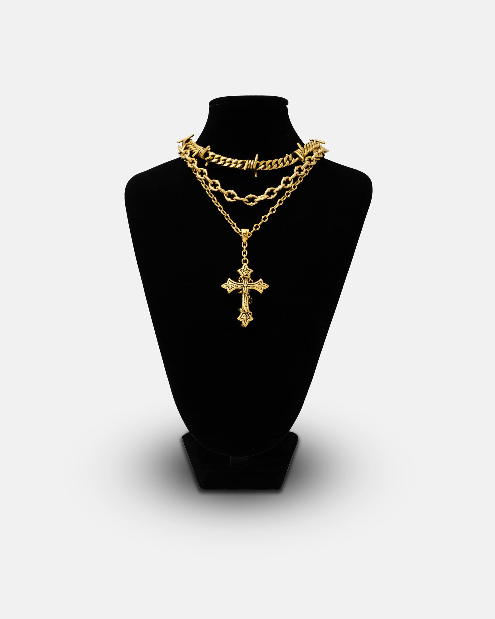 The Spite Stack Gold (Custom Pendant Choice) features layered gold chains, including 18k gold and a spiked chain, displayed on a black velvet jewelry bust. The longest chain has an ornate cross pendant against a plain light gray background.