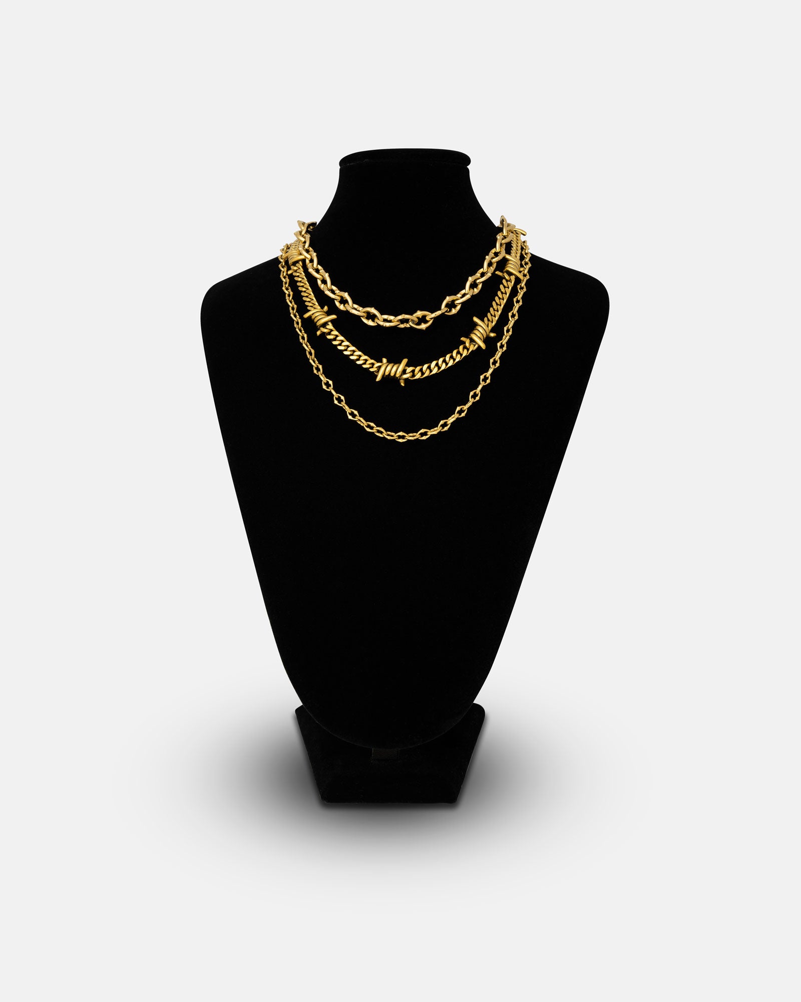 The Stay Safe Stack (Gold) features three layered gold chains in varying styles, including a bold barbed wire necklace, displayed on a black jewelry bust against a plain white background.
