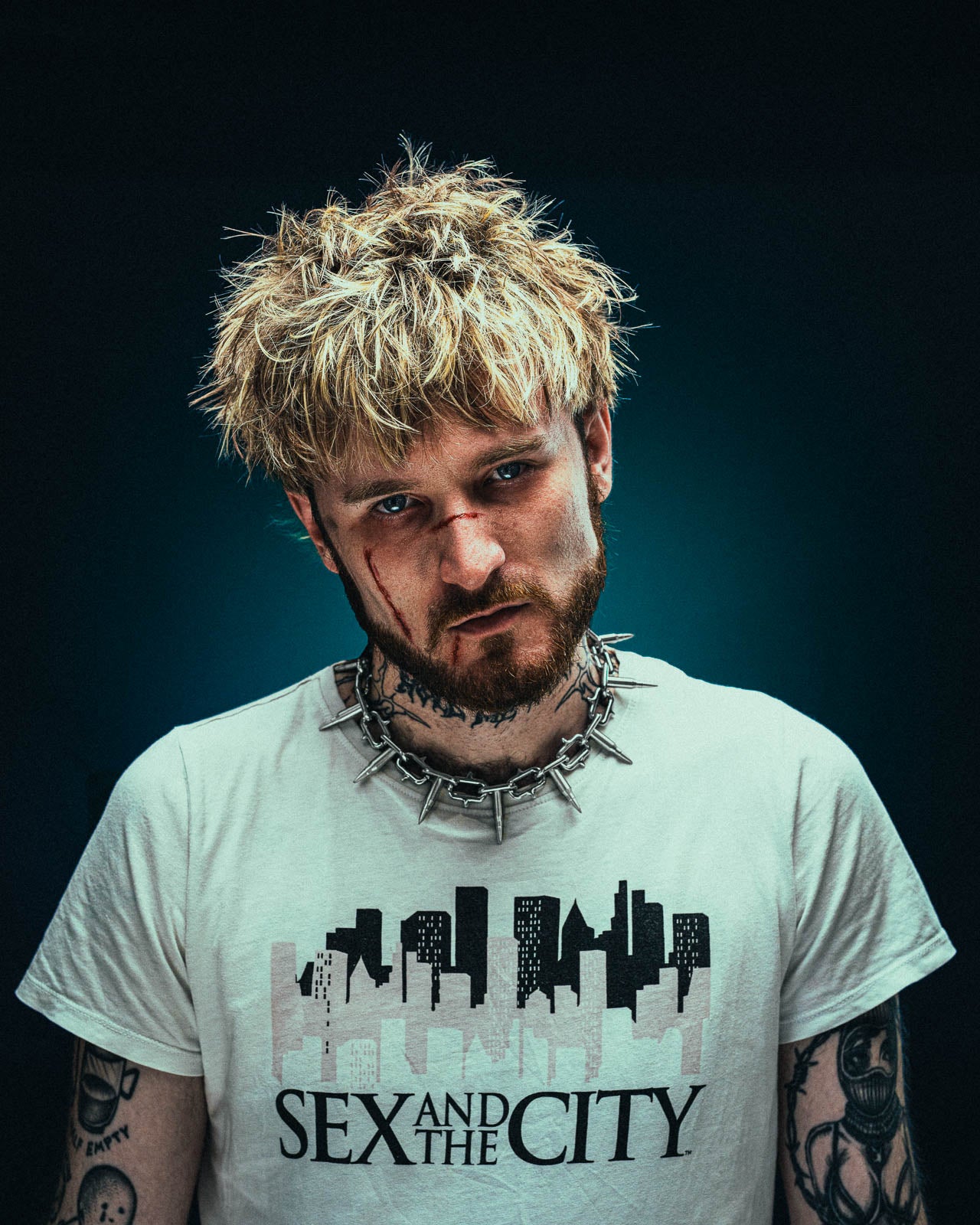 A man with messy blond hair, facial bruises, and tattoos wears a Sex and the City T-shirt and a Statement Collective "Sweating Bullets" Necklace Chain, looking seriously at the camera against a dark background.