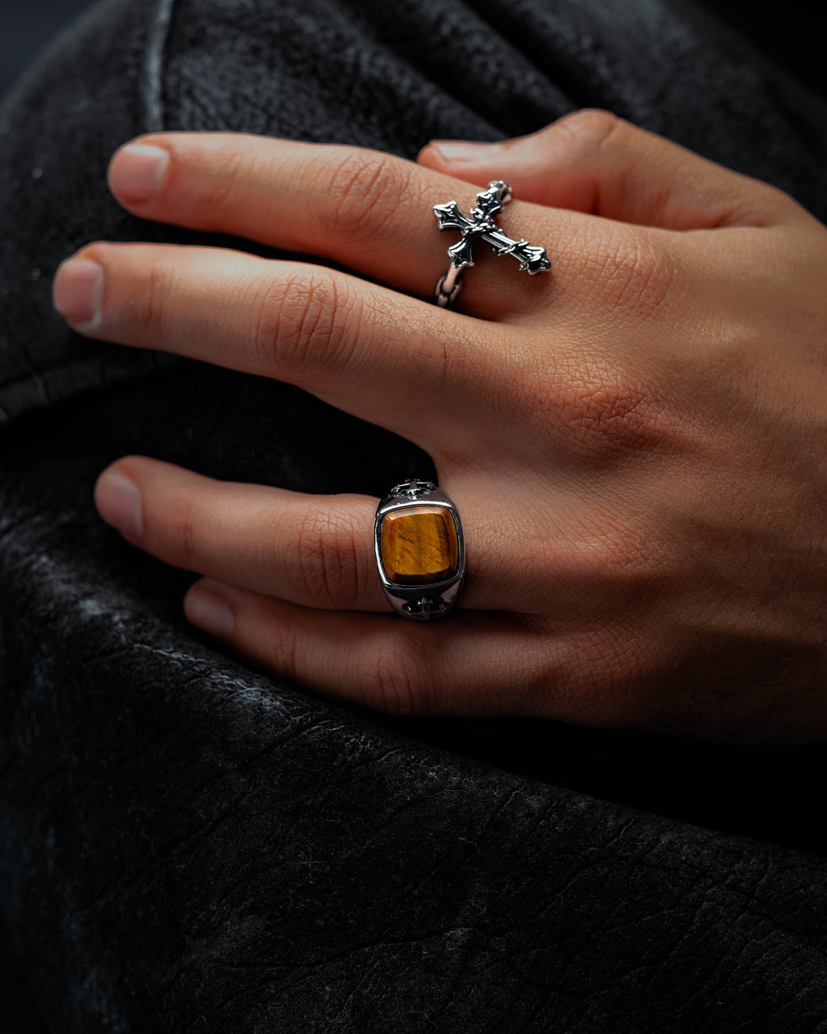 A hand adorned with a silver graveyard ring with a cross and the Statement Collective Tigers Eye Stone Ring—a stainless steel band featuring a rectangular tiger’s eye stone—rests on a black leather surface.