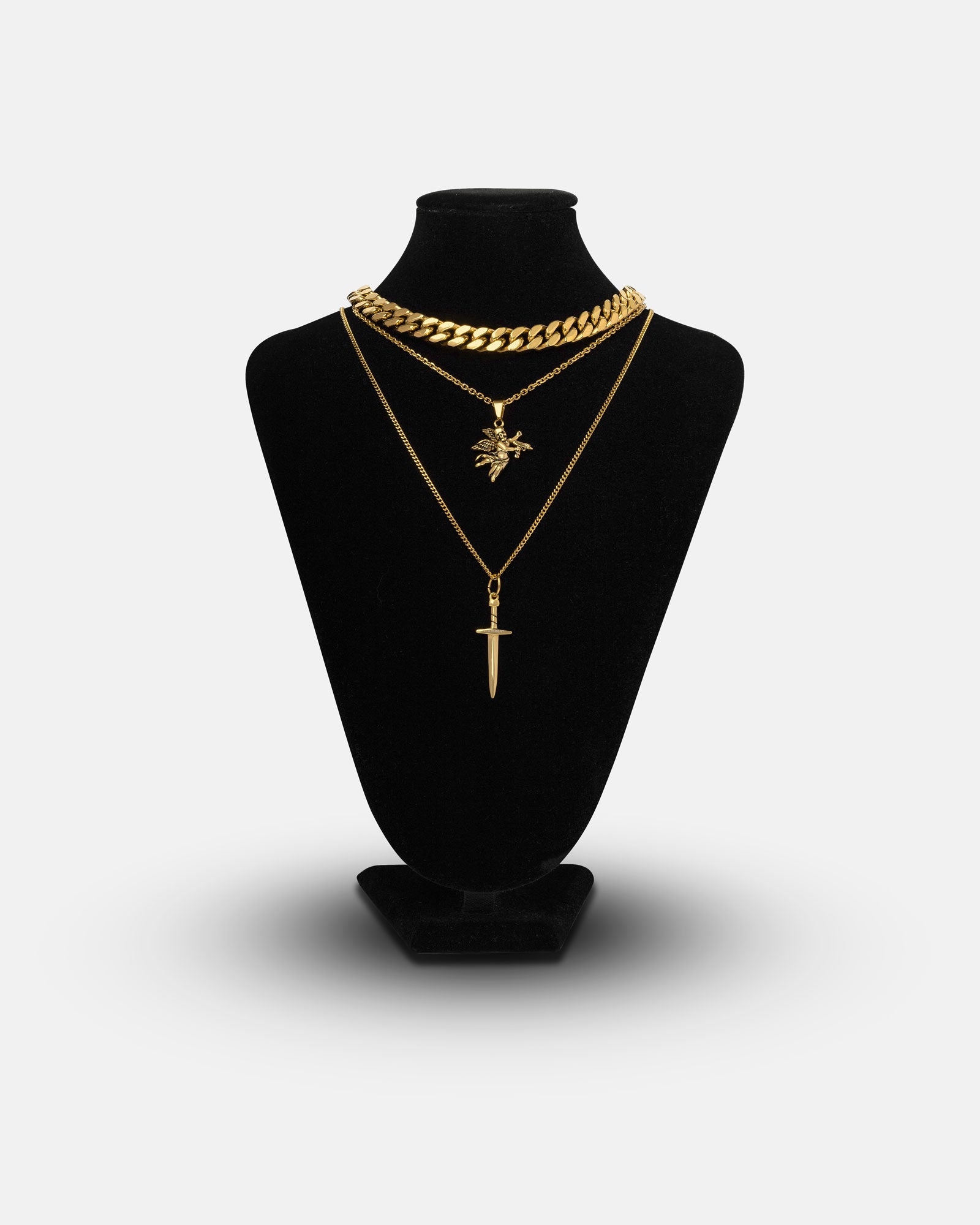 The Angel & Dagger Stack (Gold) is displayed on a black velvet stand, featuring a bold Cuban link necklace, a chain with spiked star pendant, and a longer sword pendant, all set against a plain white background.