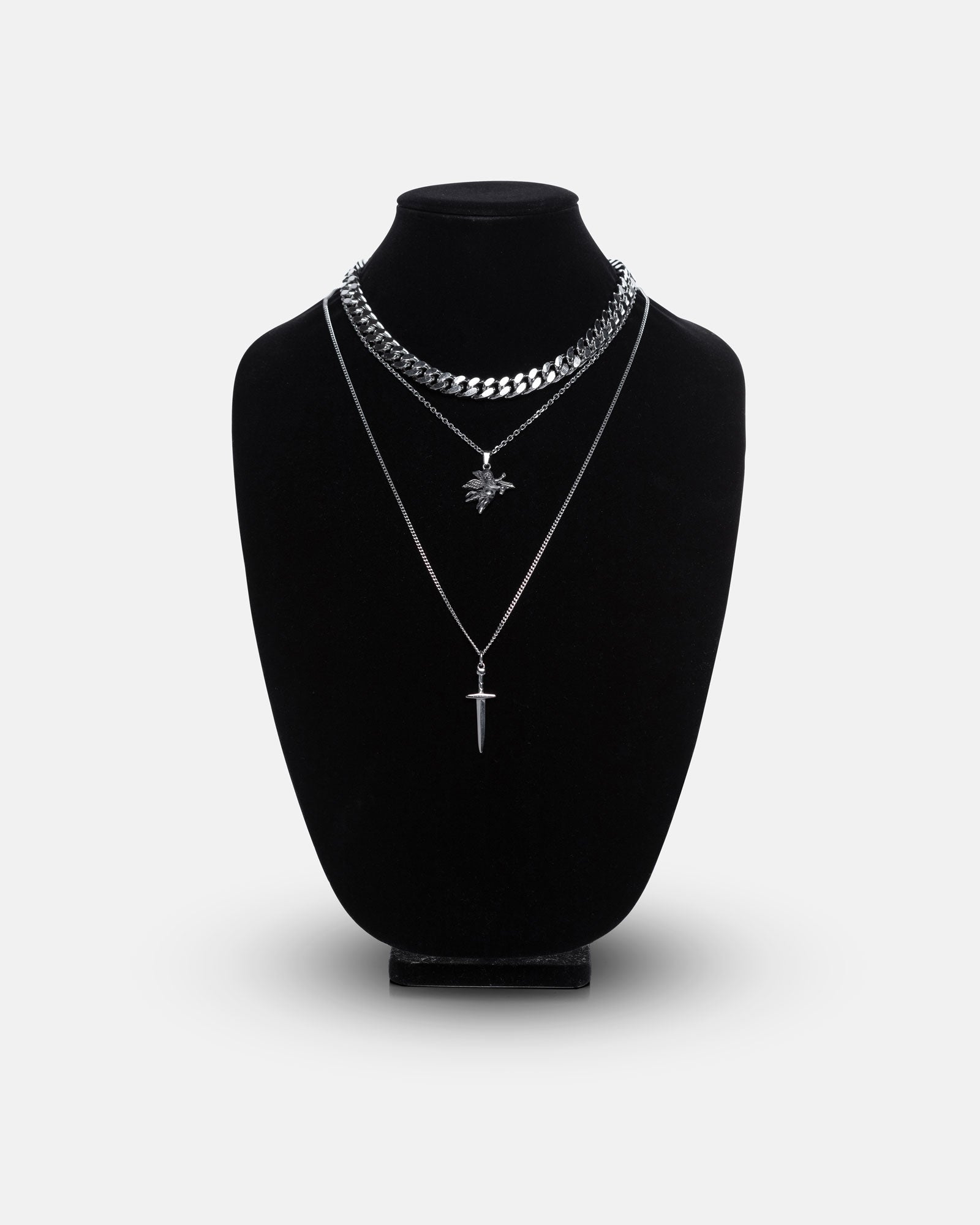 The Angel & Dagger Stack features three layered silver necklaces—a bold Cuban link, a leaf pendant, and a thin dagger-shaped chain—on a black velvet jewelry bust set against a plain white background.