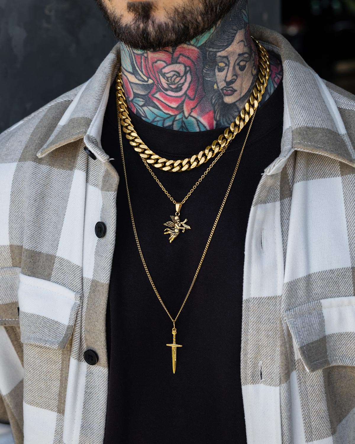A person wearing a beige and white plaid shirt over a black tee, showcasing the Angel & Dagger Stack (Gold) by Statement Collective and a colorful neck tattoo with a rose and woman's face.