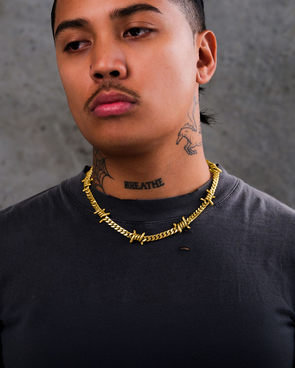 A person wearing a dark gray shirt, the 6mm Barbed Wire Cuban Chain (Gold), and visible neck tattoos including the word BREATHE. The background is a plain gray surface.