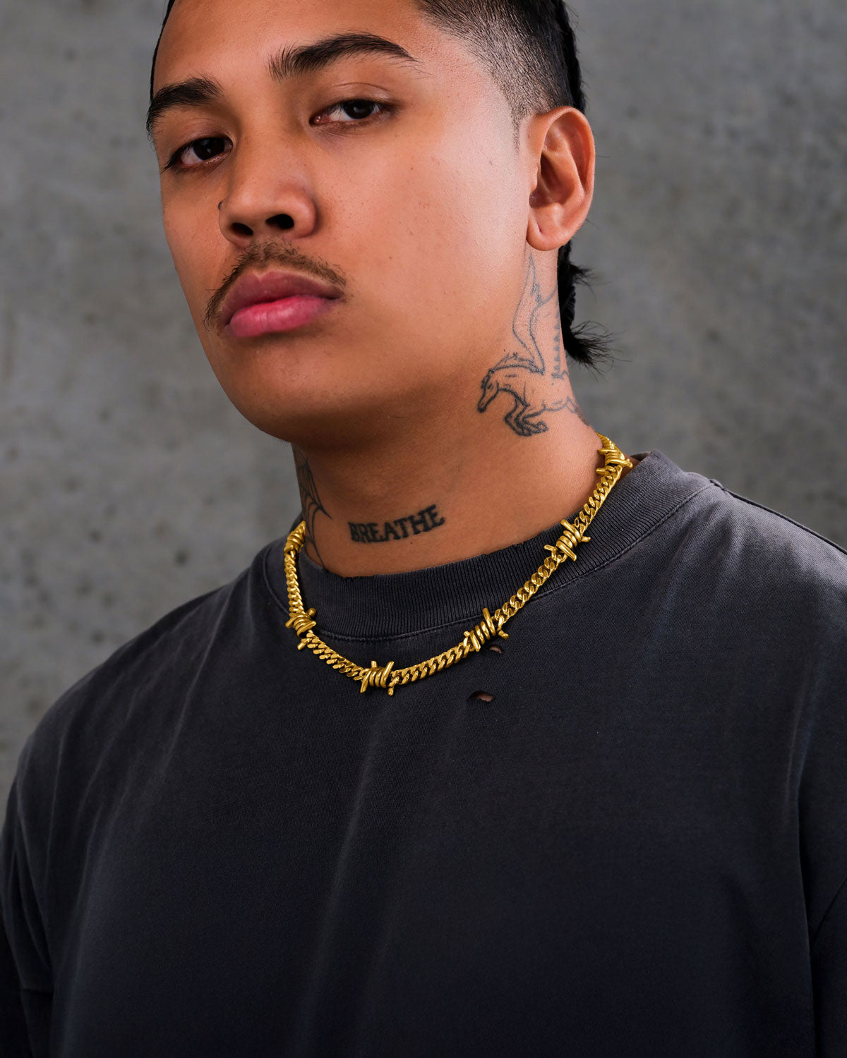 A person with a mustache and a "BREATHE" neck tattoo wears a dark t-shirt and the 6mm Barbed Wire Cuban Chain (Gold), standing against a gray textured background.
