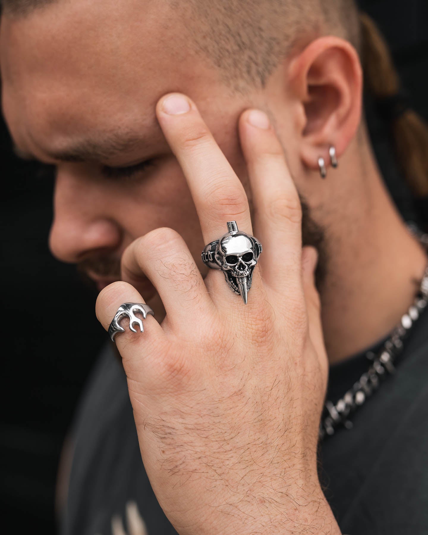A person with a beard, earrings, and a side-shaved hairstyle holds up two fingers near their face, showcasing bold rock jewelry—a "Bite The Bullet" skull ring and a gothic flame ring. They also wear a chain necklace.