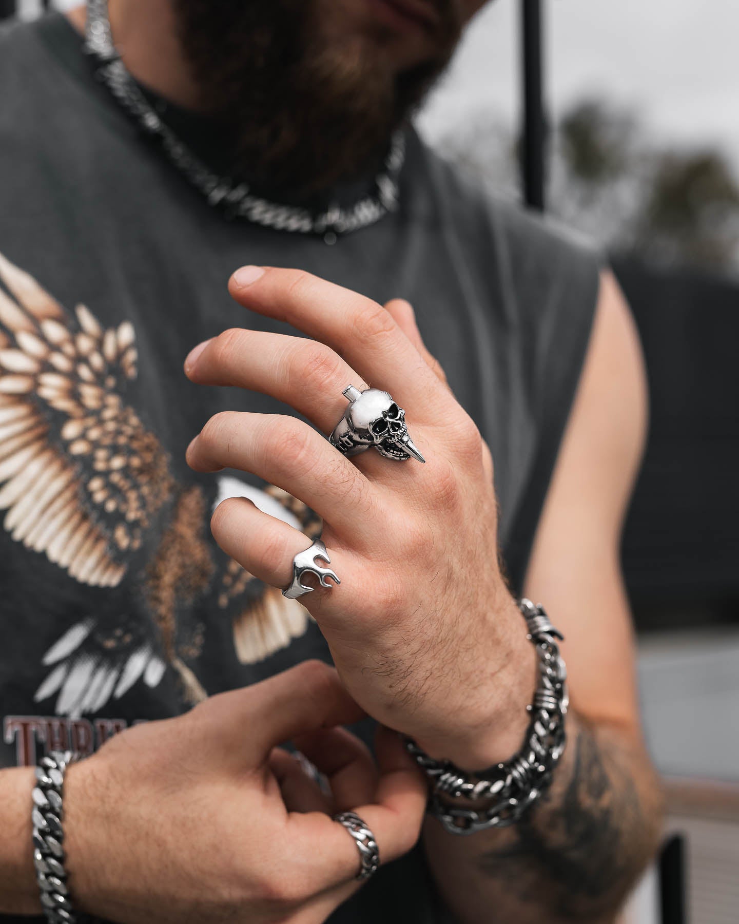 A person in a sleeveless eagle-print shirt flaunts rock jewelry—silver rings, chunky chain bracelets, and the "Bite The Bullet" Ring—while their bearded face is partly visible against a blurred background.