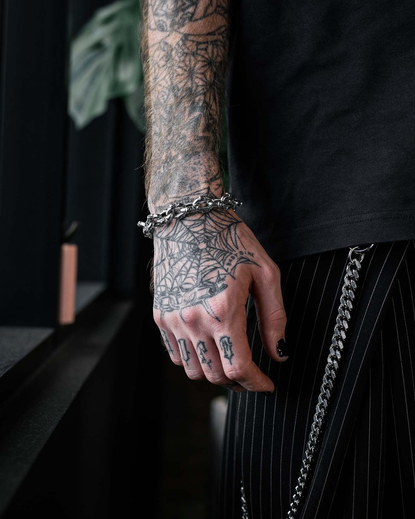 A tattooed hand adorned with the "Boneyards" Skull Bracelet by Statement Collective and rings rests by the side, wearing black pinstripe pants with chain accessories. The person sports black nail polish and a web tattoo on their hand.