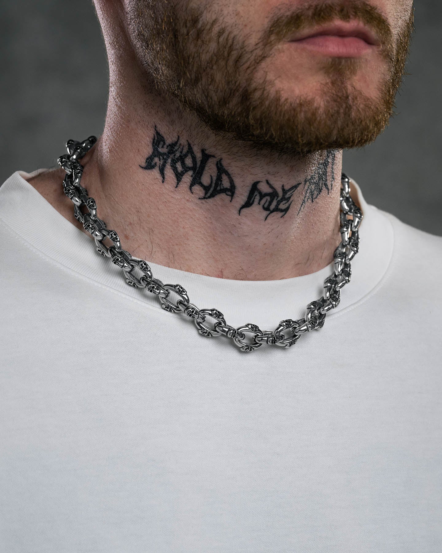 A close-up of a man wearing the "Boneyards" Skull Chain necklace and a white shirt, showing a neck tattoo that reads “HOLD ME TIGHT” in bold, stylized letters.