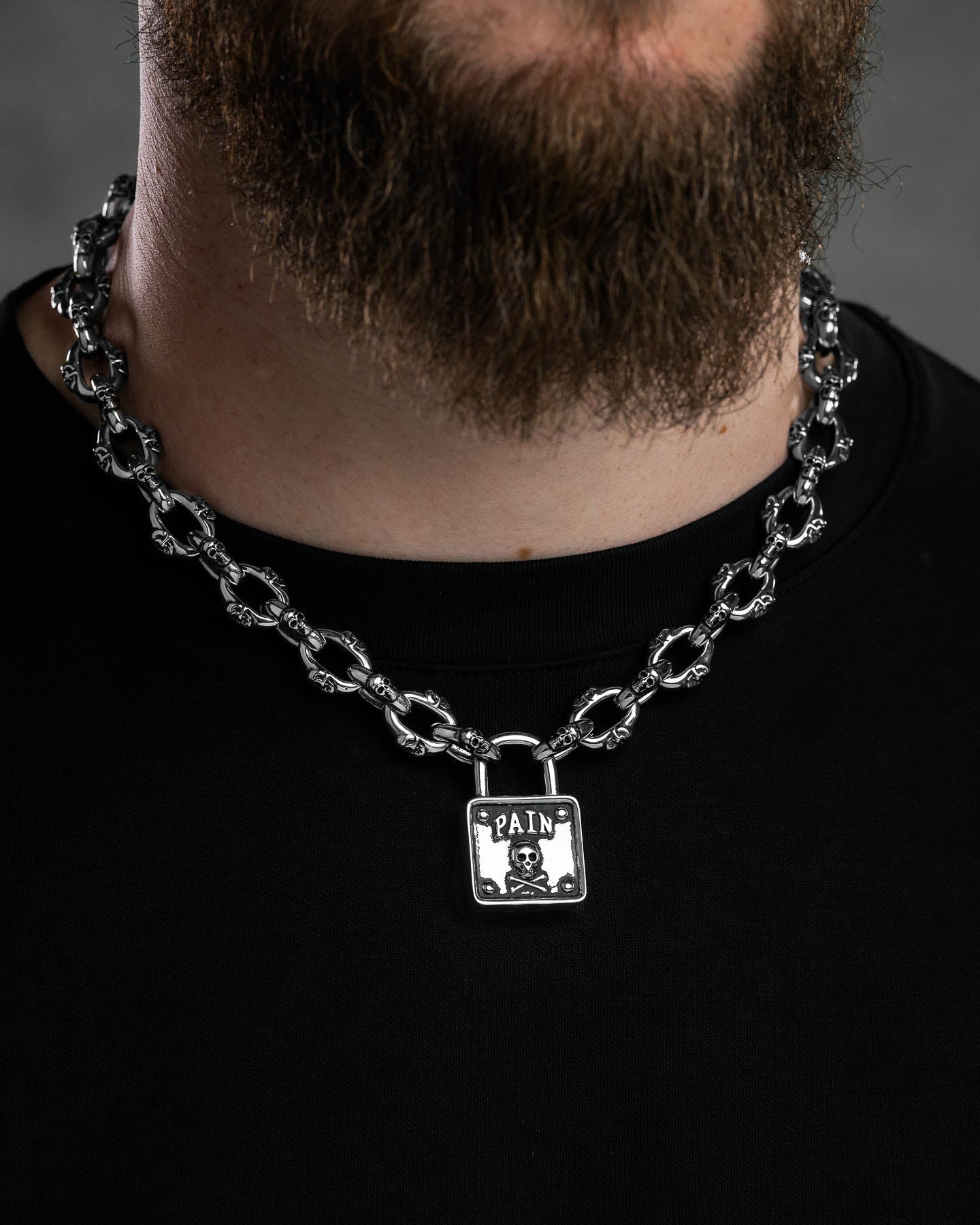 A bearded person wears a black shirt and the "Boneyards" Skull Padlock Chain, a bold 12mm stainless steel necklace with a square pendant reading PAIN above a skull and crossbones.