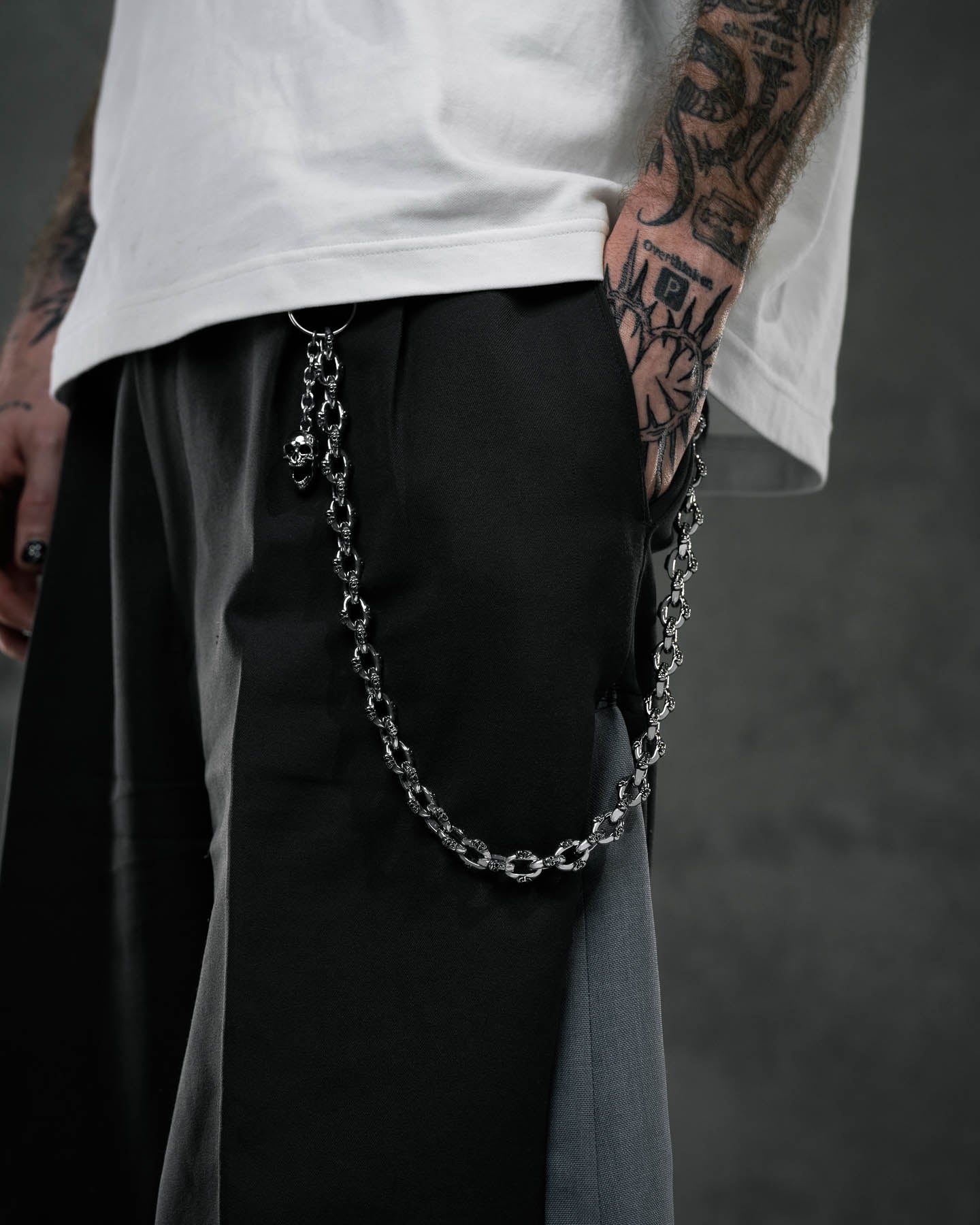 A person wearing black and gray pants with the "Boneyards" Skull Wallet Chain clipped to the pocket stands with a tattooed hand in the pocket, a partially visible white shirt, and a dark, blurred background.