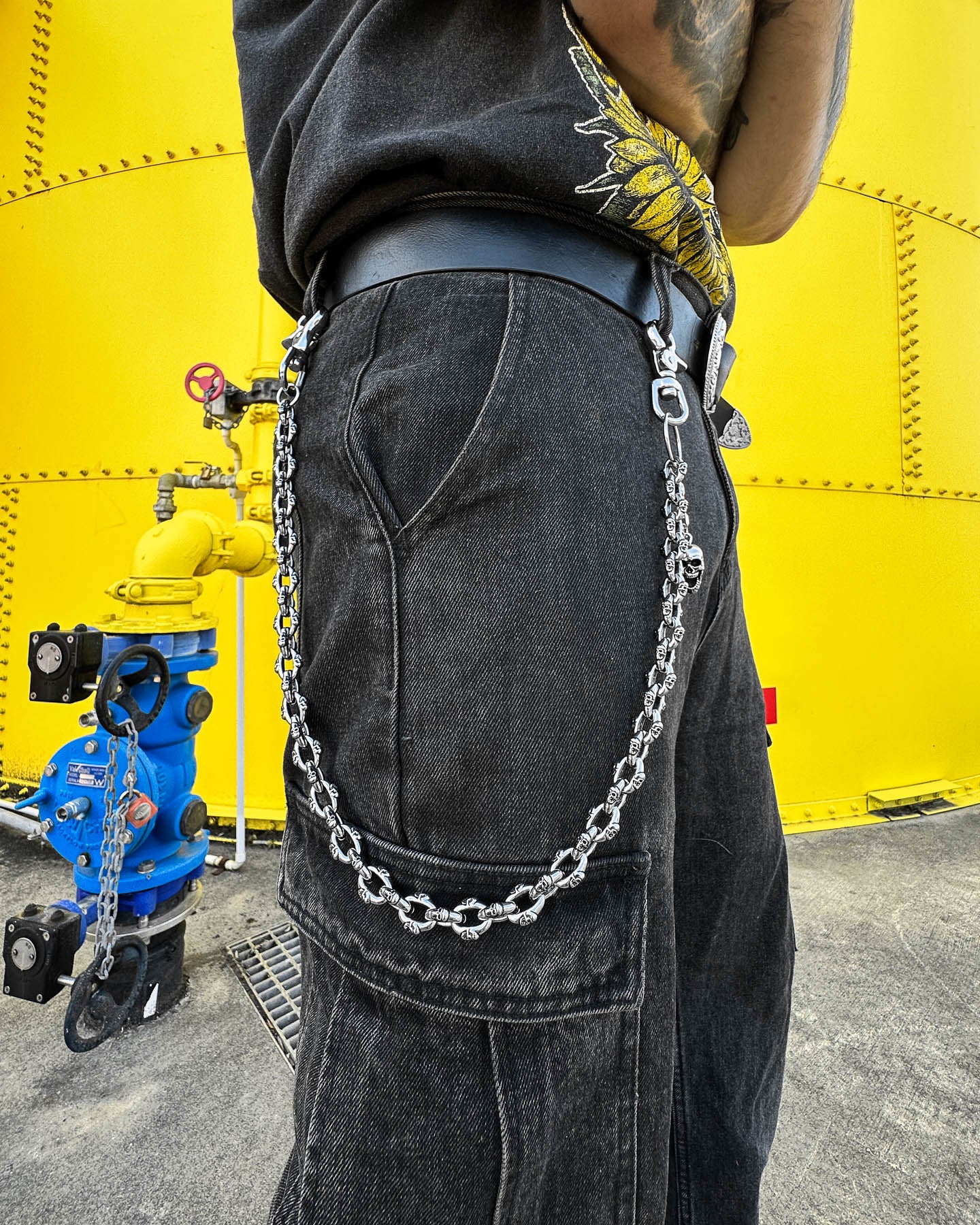 A person wears black pants with the "Boneyards" Skull Wallet Chain clipped to a belt loop, standing by a bright yellow industrial tank and blue fire hydrant. Only their lower torso and hip are visible.