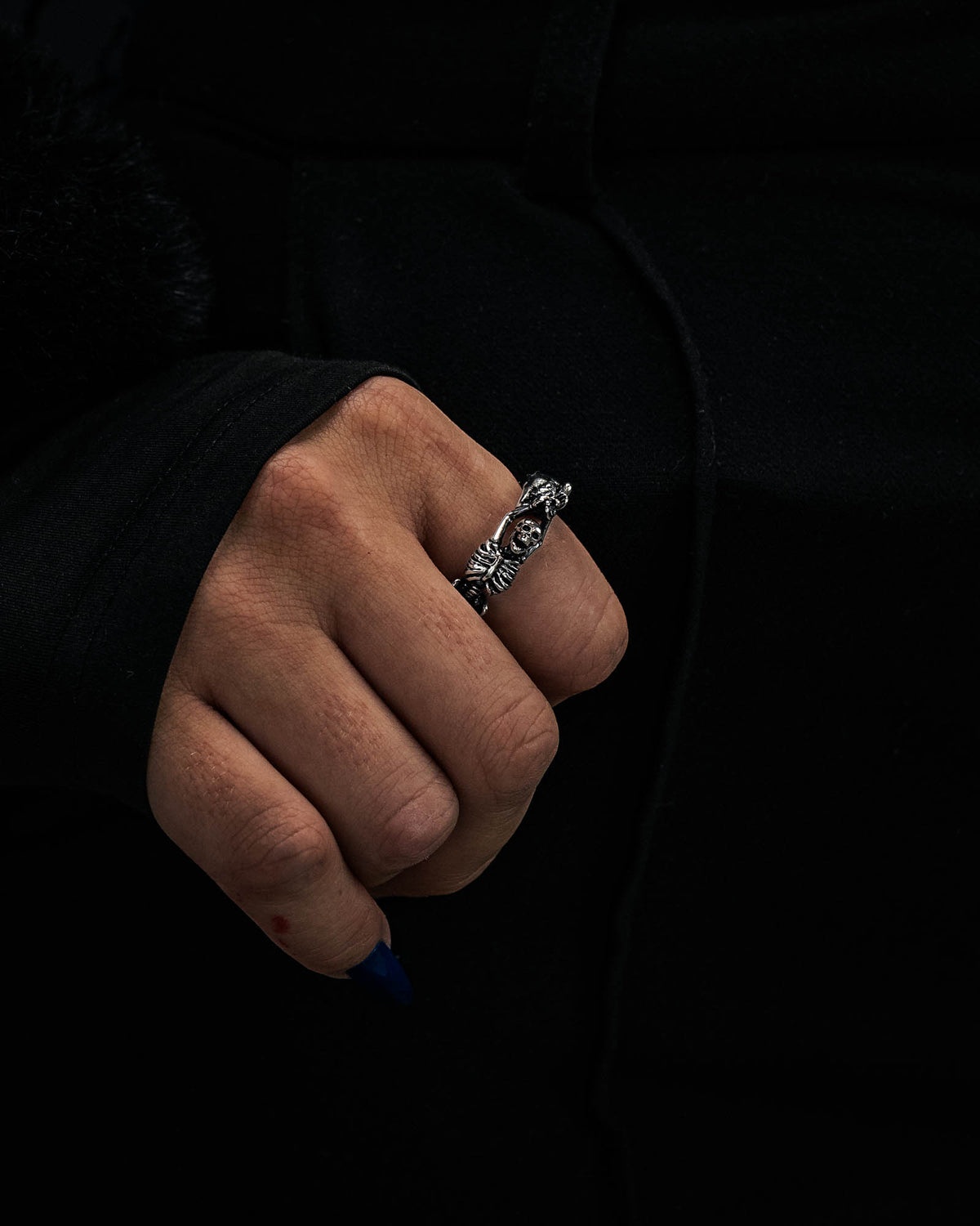 A hand with dark blue nails showcases the Statement Collective Breaking Wheel Ring— a silver, textured piece featuring sculpted faces—against a dark background, with the wearer dressed in black clothing.