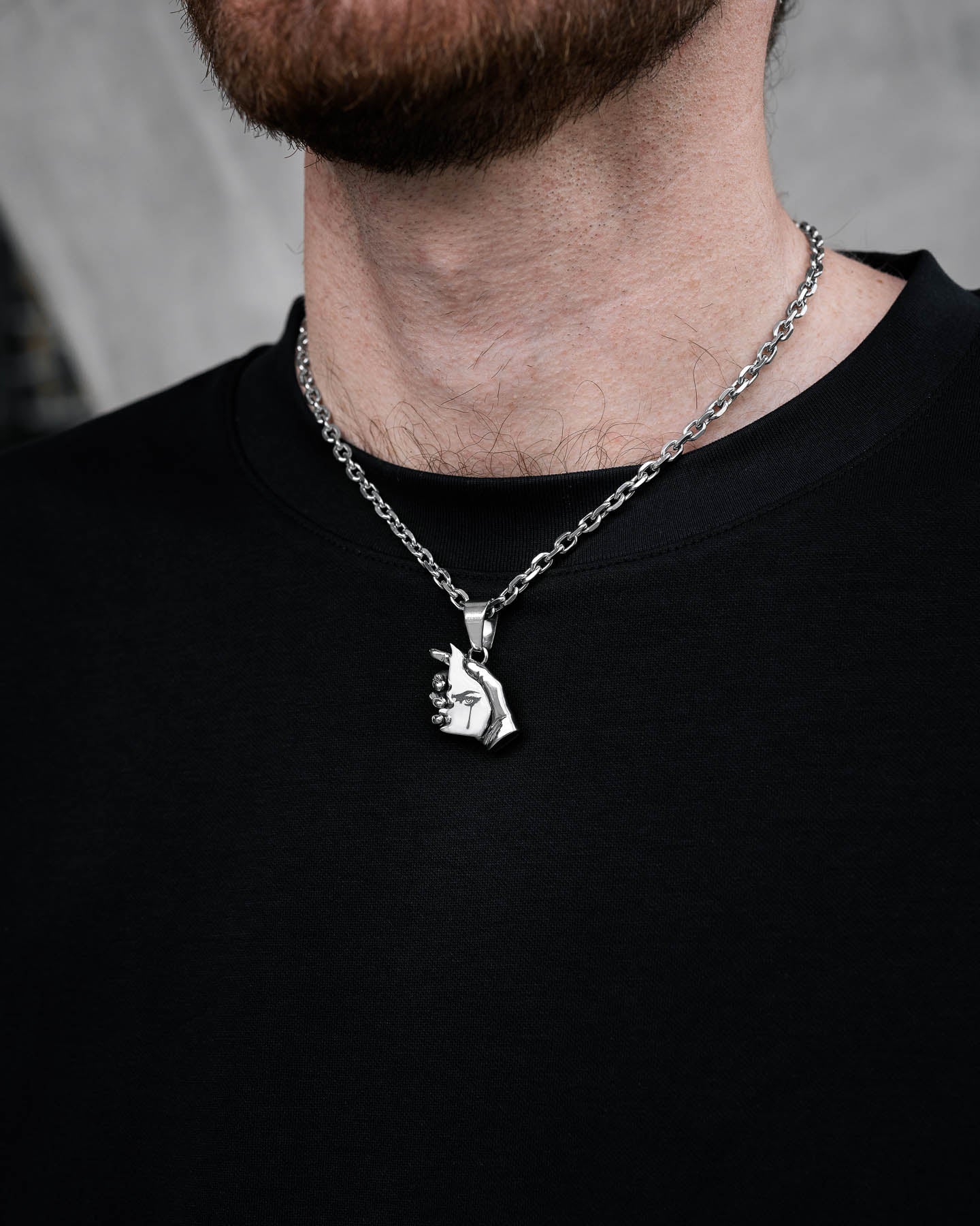 A person wears "The Broken Mirror" Pendant—a silver chain necklace with a hand holding a face pendant—paired with a plain black shirt. The image highlights the neck and chest area.