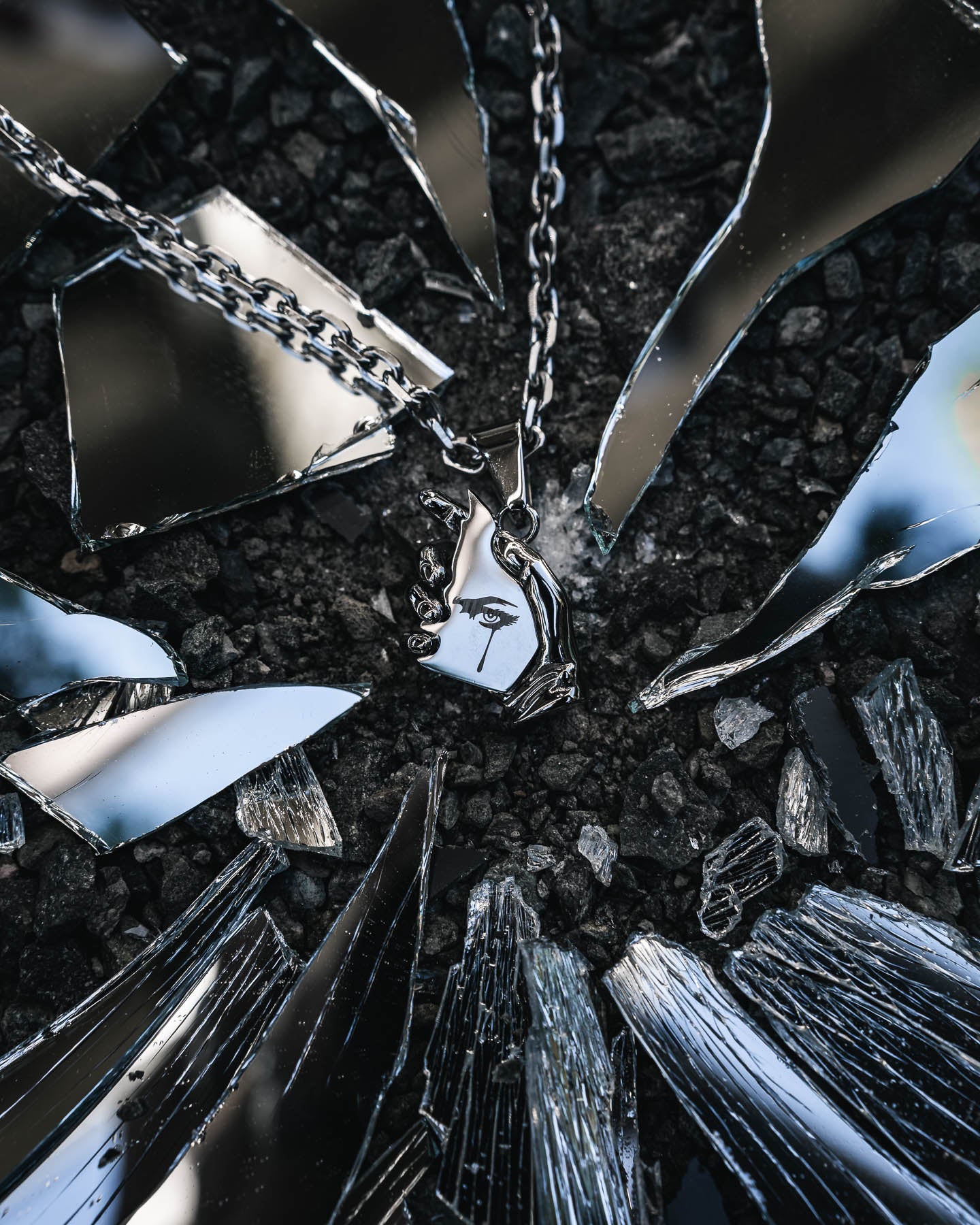 The Broken Mirror" Pendant, shaped like a stylized face and crafted in gleaming 316L stainless steel, rests on dark gravel amid mirror shards that reflect the sky—an avant-garde silver jewelry statement.