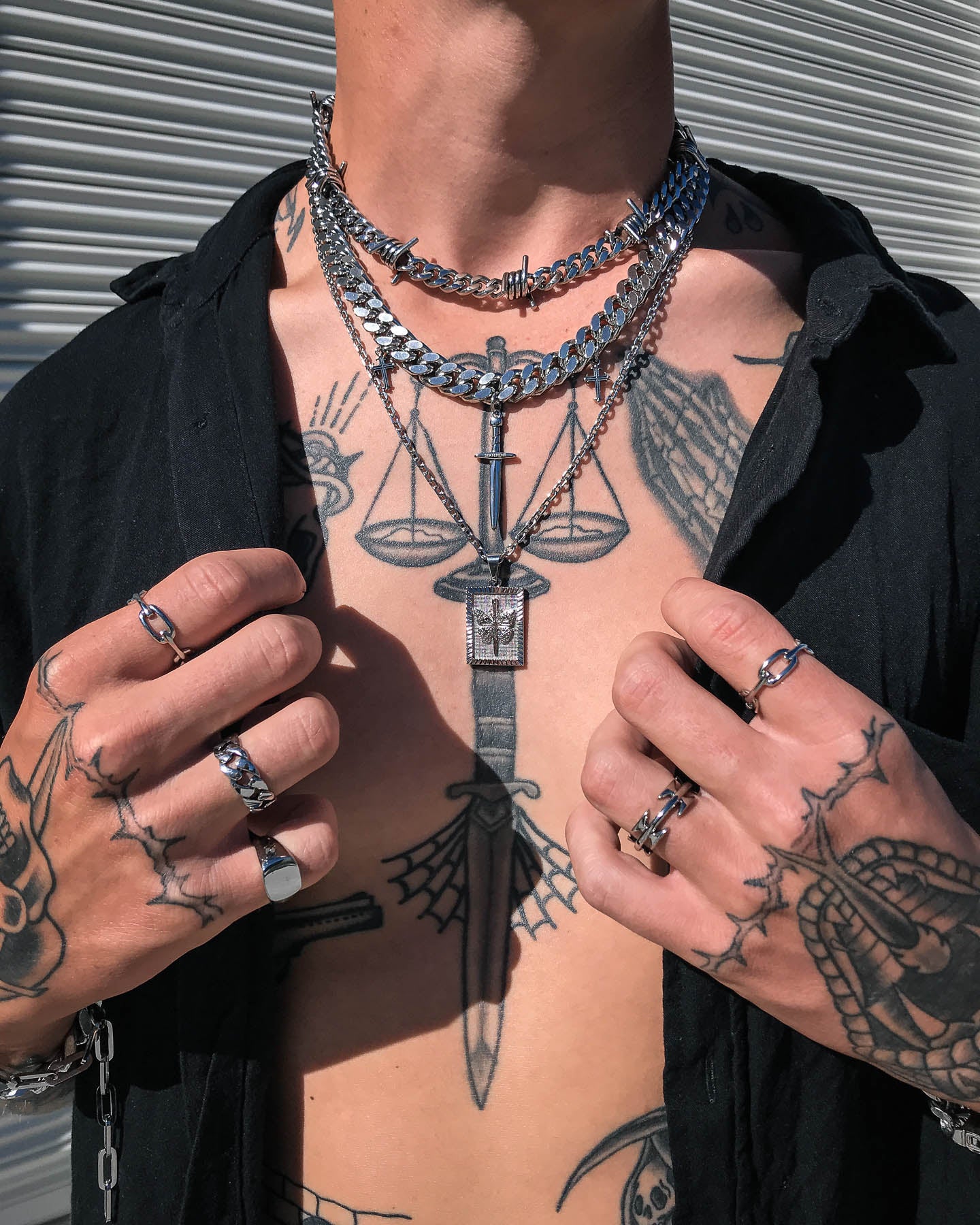 A person with tattoos on their chest and hands wears layered silver necklaces, including a Butterfly Tag Pendant, plus chunky rings. An open black shirt reveals the detailed body art underneath.