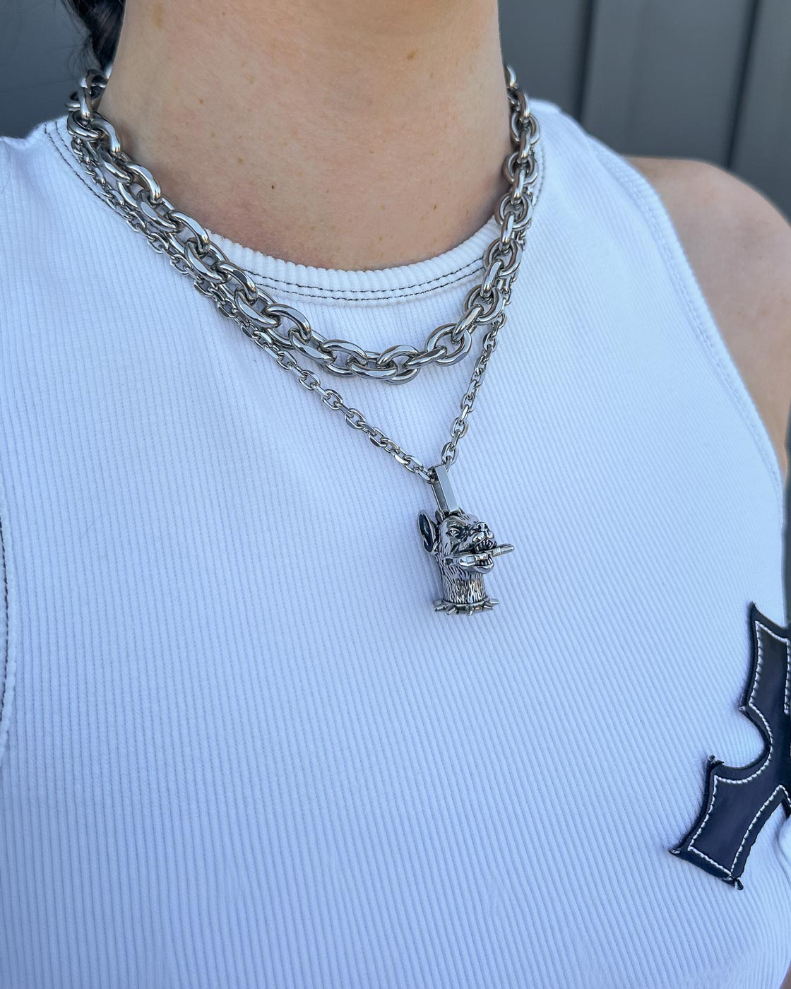 A person wears a white ribbed tank top with a black patch and two chunky stainless steel chain necklaces from Statement Collective’s Cable Set (Custom Pendant Choice), including one with a small devil character and trident pendant.