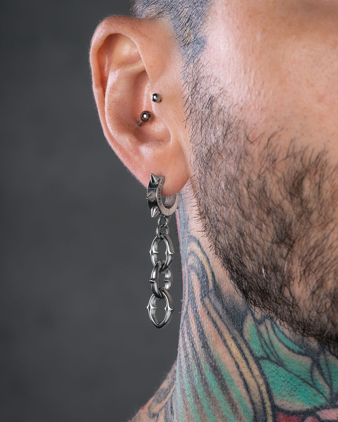 Close-up of a person’s ear wearing "The Cathedral" 9mm Chain Earrings, featuring a black stud in the upper cartilage and a detailed, colorful neck tattoo. The gray background is blurred.