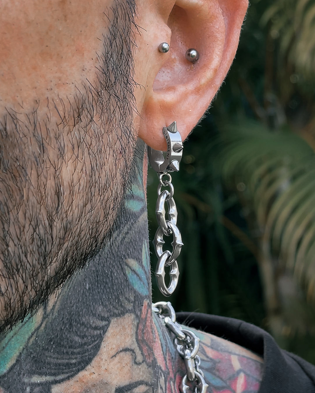 Close-up of a bearded person with neck tattoos wearing two silver piercings and "The Cathedral" 9mm Chain Earrings. Blurred greenery is visible in the background.