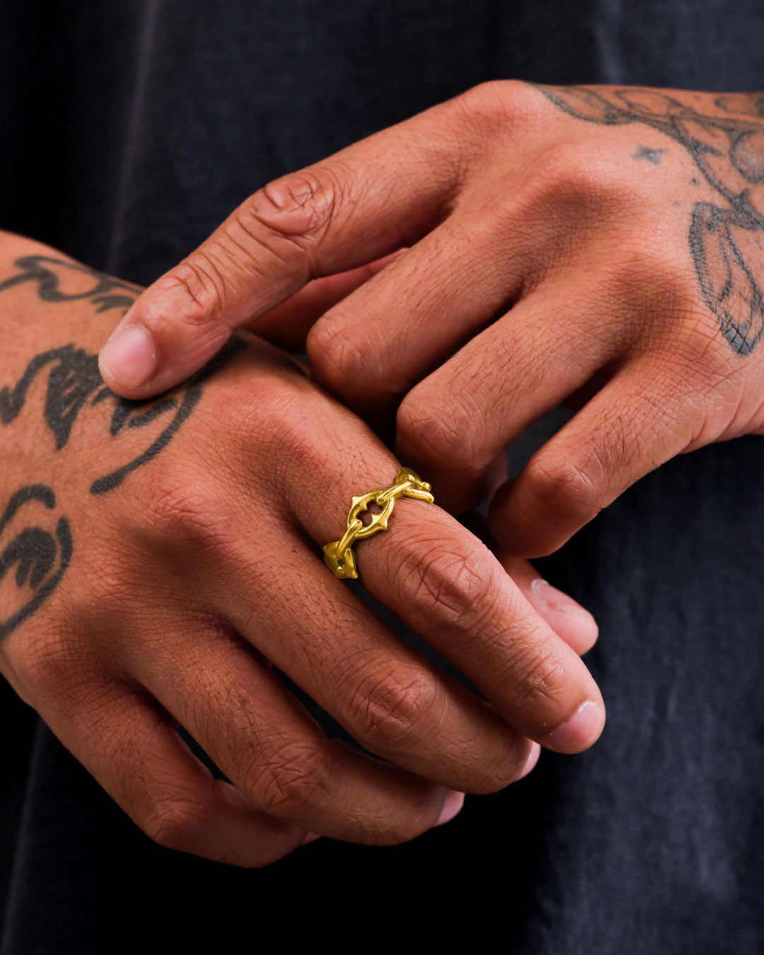 A close-up of tattooed hands, one gently touching the other adorned with Statement Collective's "The Cathedral" Spiked Ring (Gold), featuring an organic, intertwined design, set against a softly blurred dark background.
